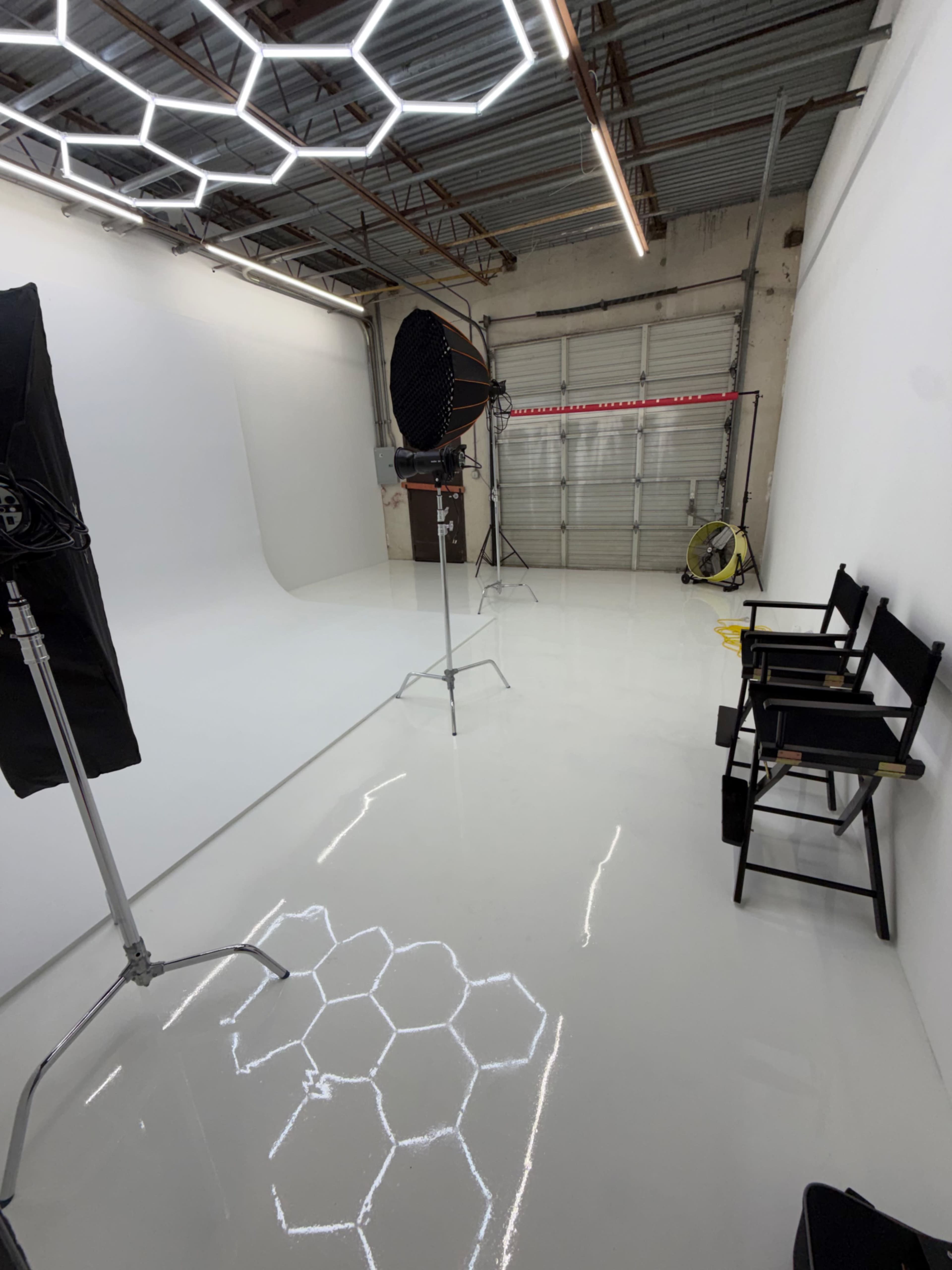 The image shows a photography studio with a white backdrop, studio lights on stands, and two black director's chairs on a shiny floor featuring a hexagonal pattern.