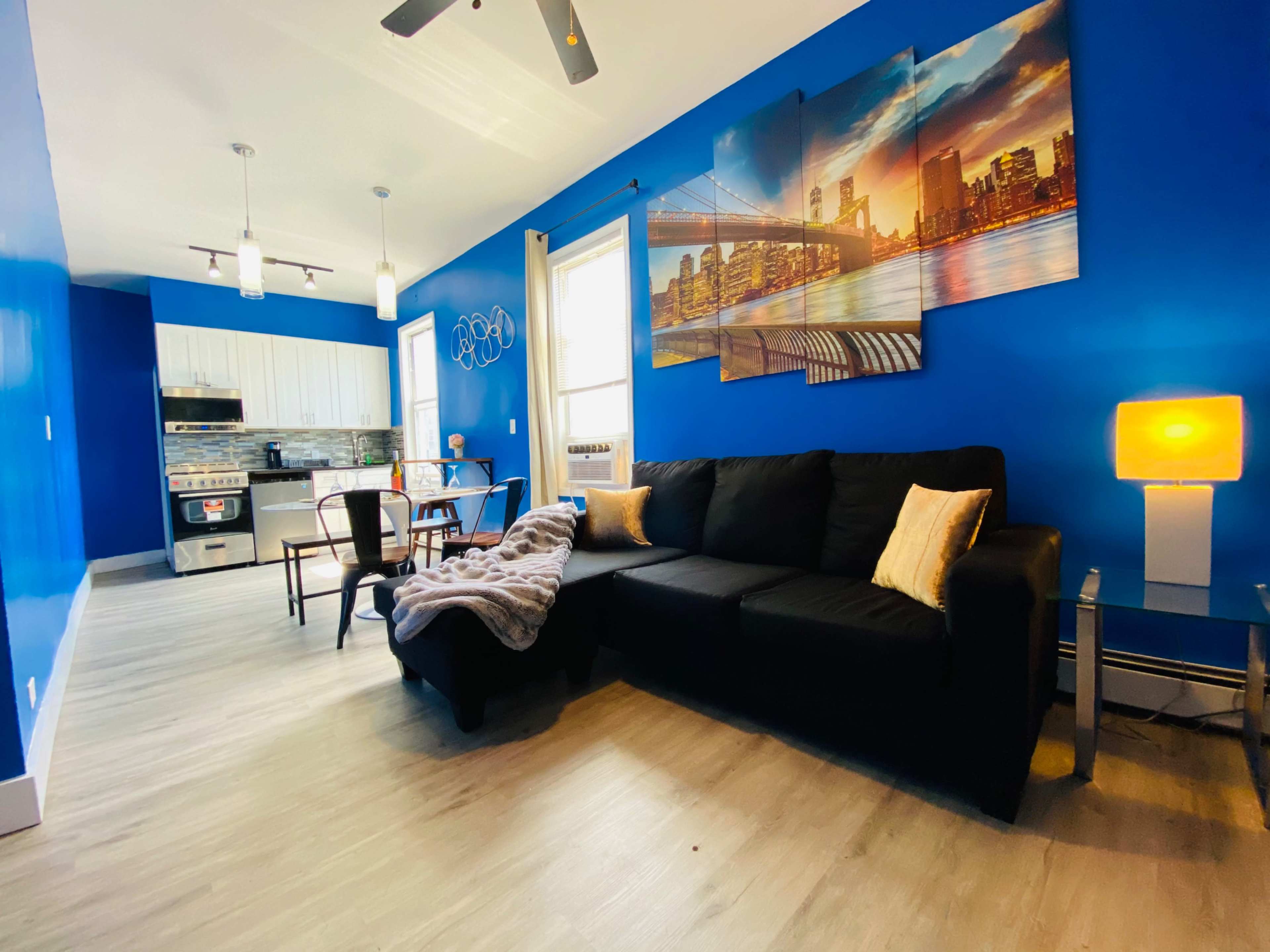 The room features a black sofa with decorative pillows, a small dining area, and a blue accent wall with a cityscape mural.