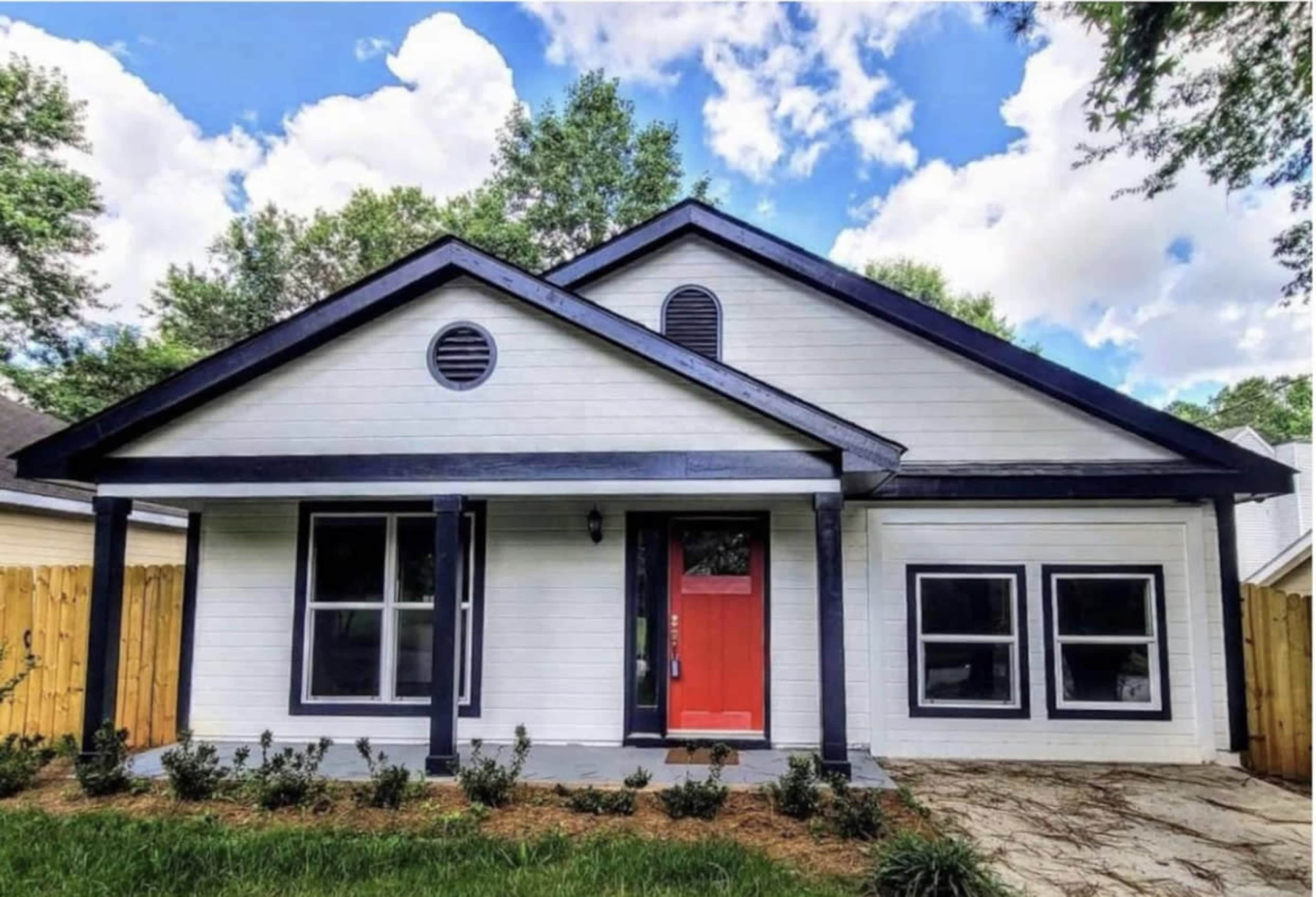A single-story house with a gable roof, white siding, and a bright red front door is surrounded by a landscaped yard and wooden fence.