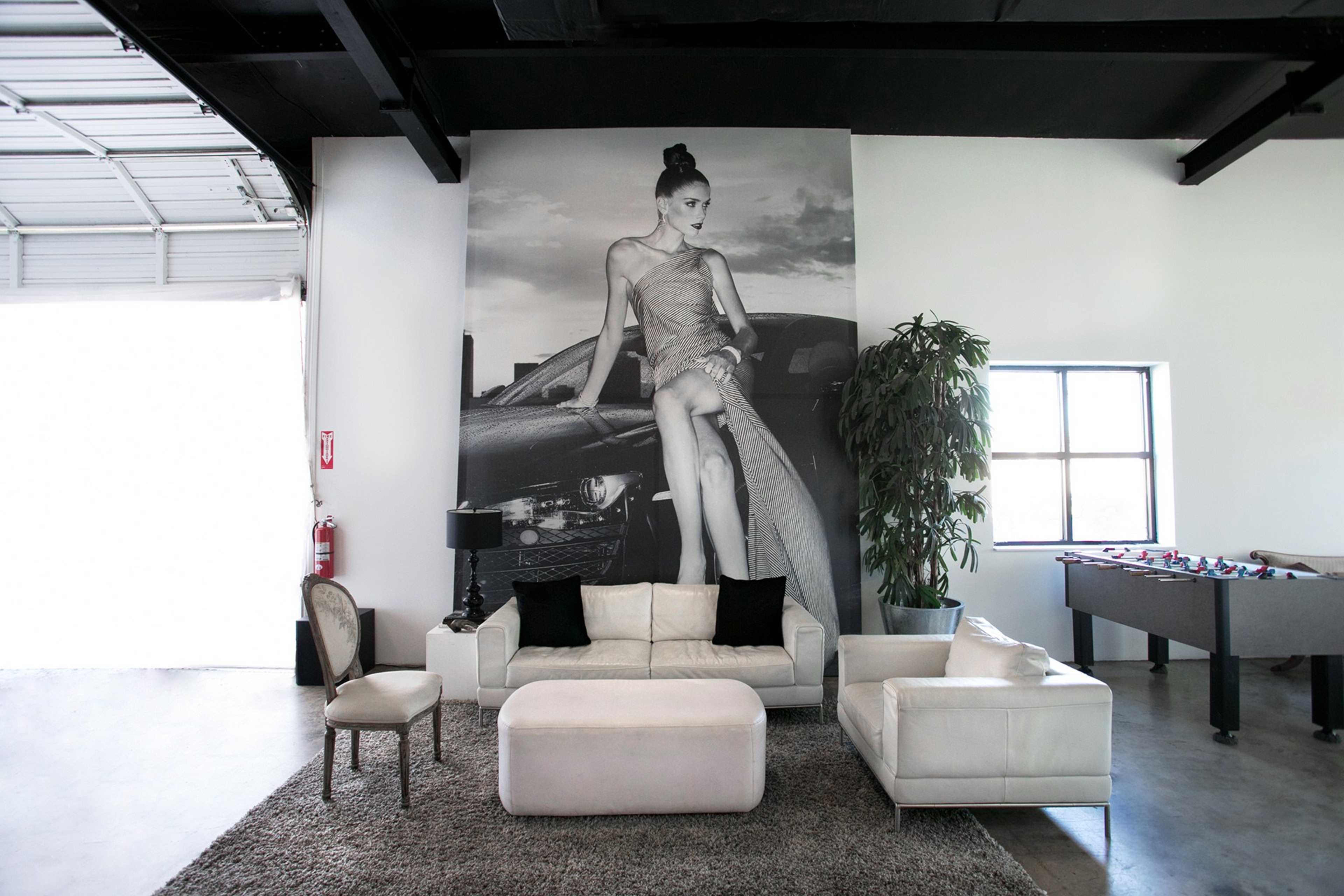 The scene features a modern lounge area with a white sofa, an ottoman, and a large black-and-white mural of a woman next to a car on the wall.