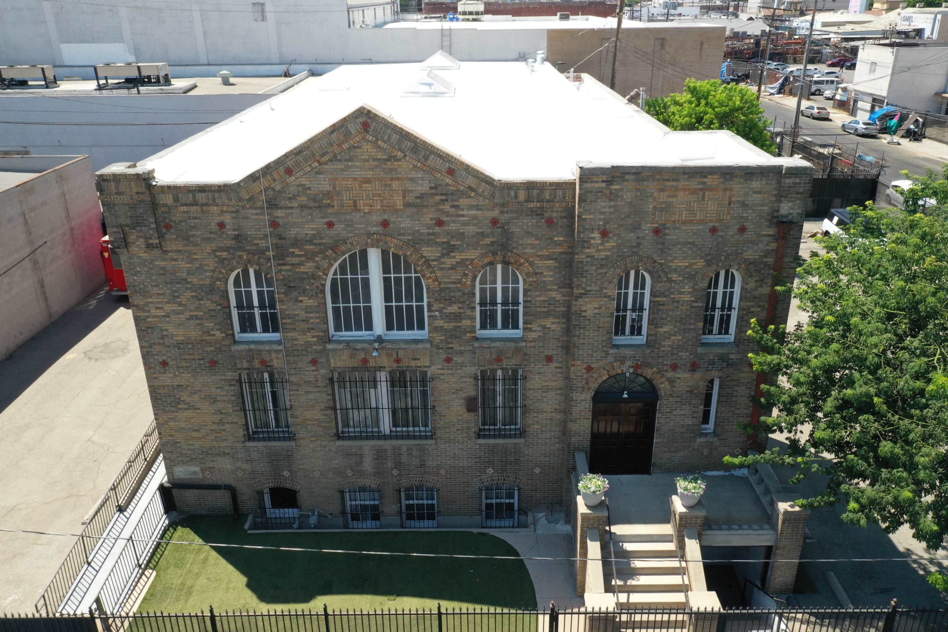 The image shows a two-story brick building with large arched windows and a prominent entrance, surrounded by a small green area and concrete pavement.