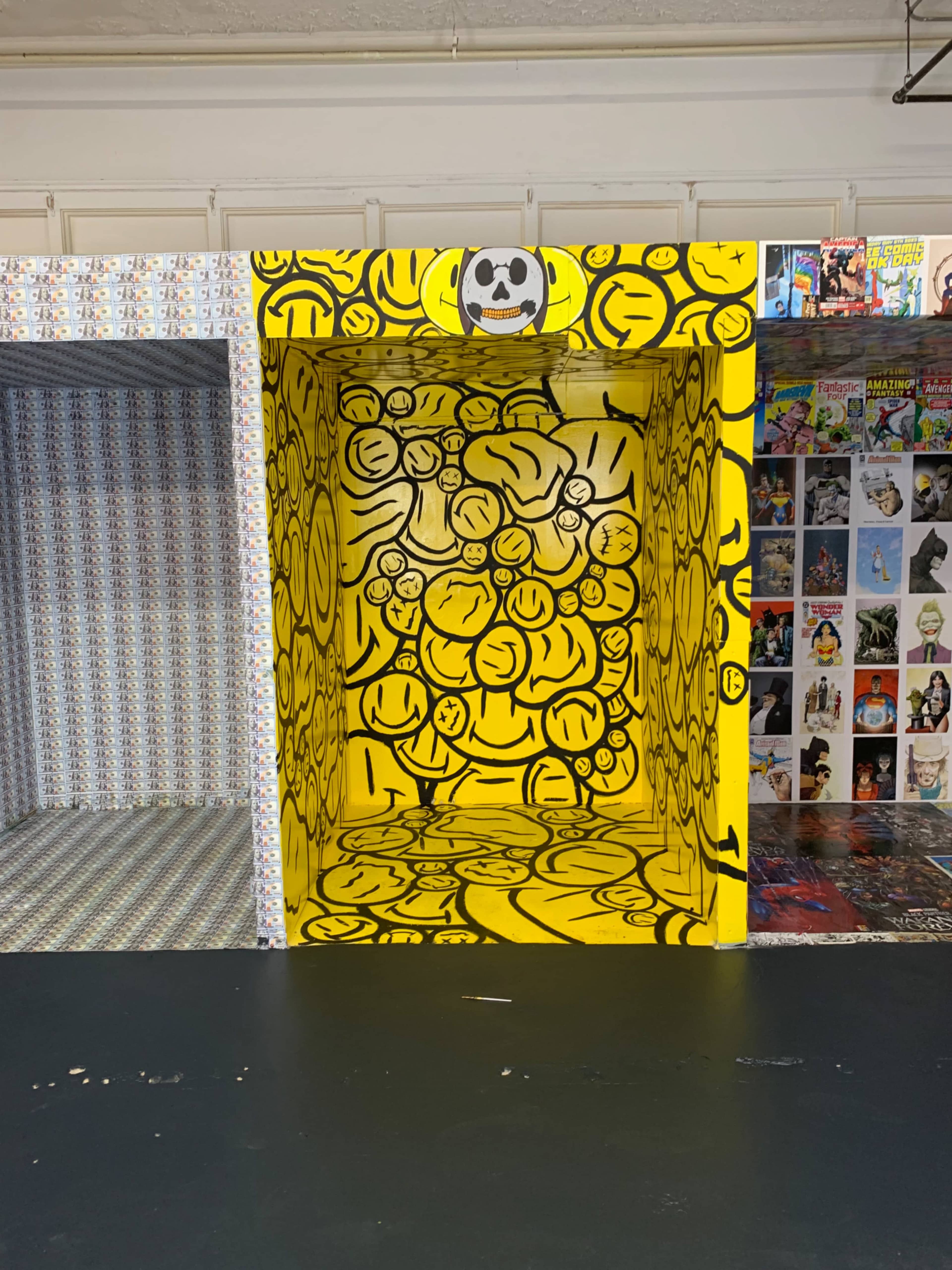 A colorful installation featuring a large yellow box with various cartoon-style smiley faces drawn on its interior walls, surrounded by patterned panels.