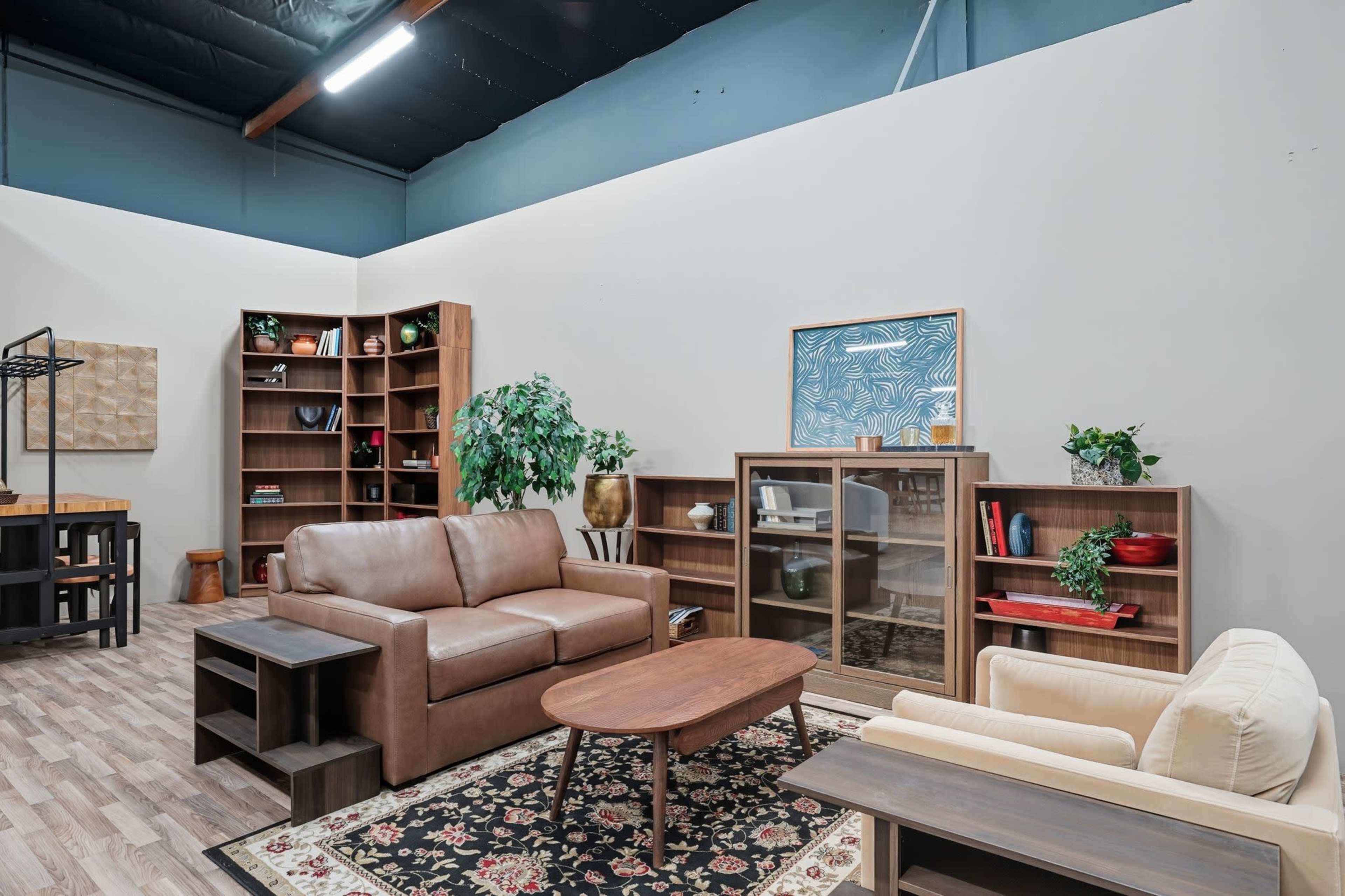 The image shows a cozy interior space featuring a brown leather sofa, a light-colored armchair, a wooden coffee table, and shelves filled with books and decorative items.