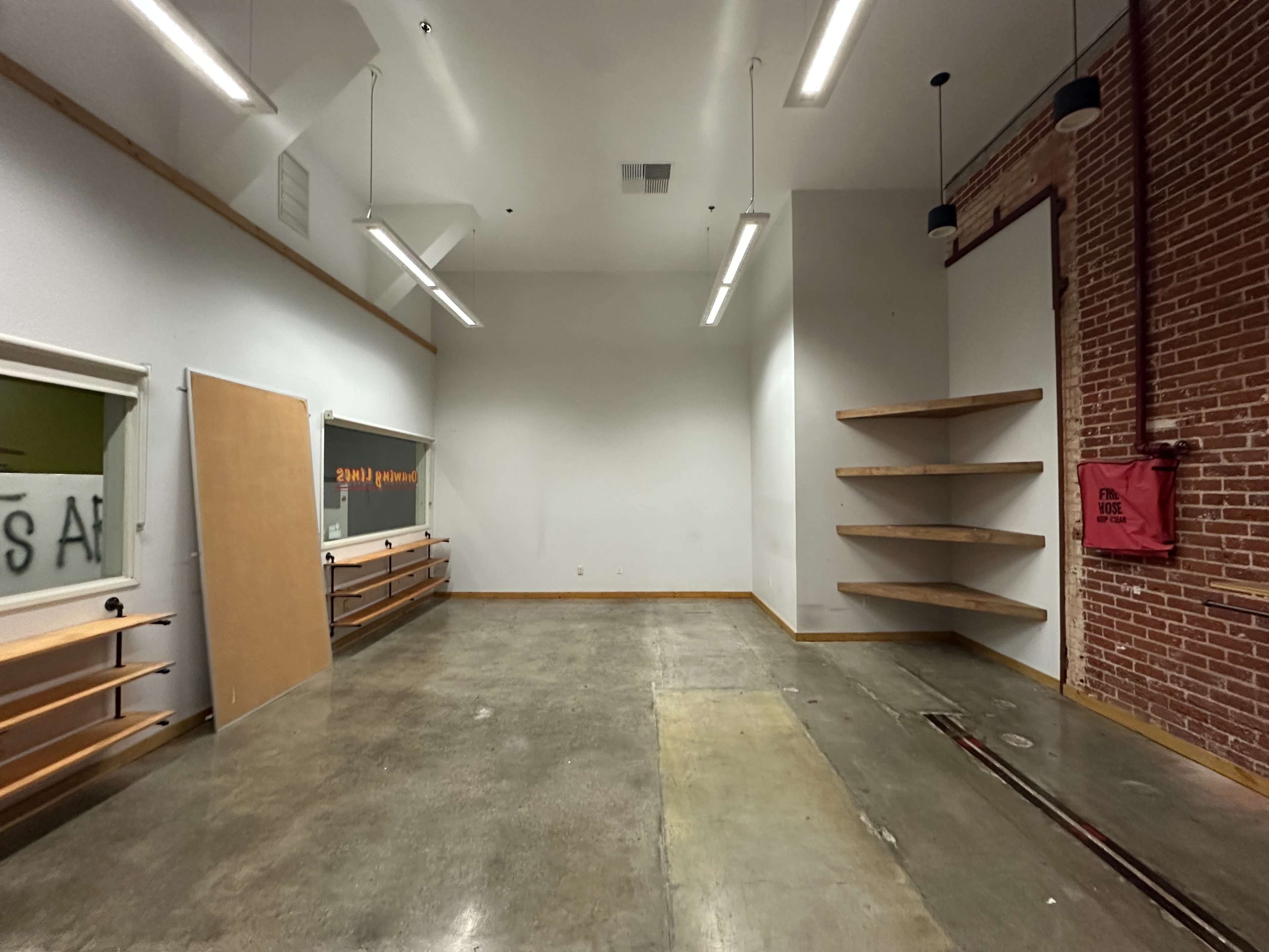 The image shows a sparsely furnished room with concrete flooring, exposed brick walls, and a few wooden shelves mounted on the side.