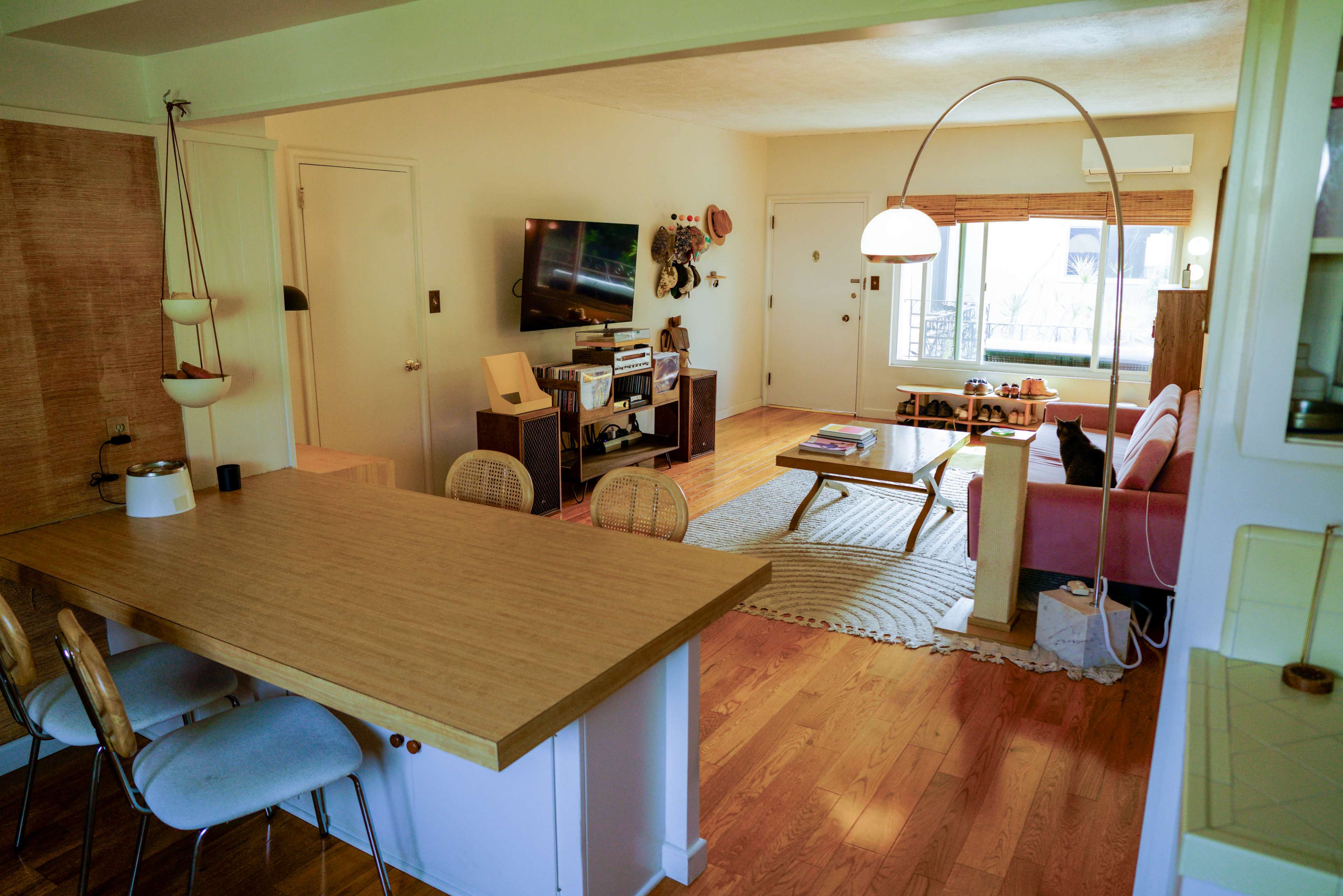 The image shows a cozy living space that features a kitchen area with wooden countertops, a living room with a pink sofa, and a large window allowing natural light to enter.