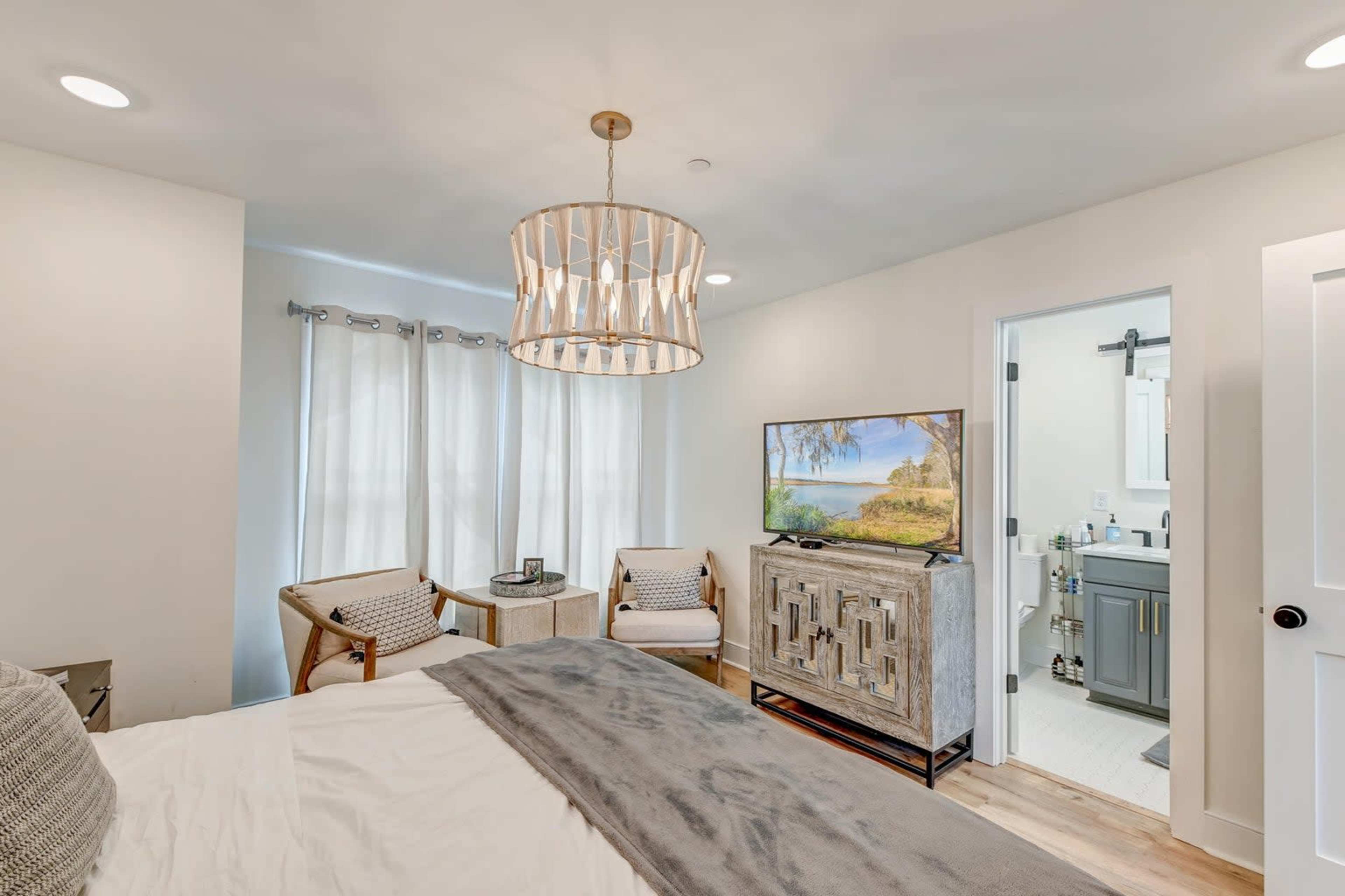 A well-lit bedroom features a bed, two armchairs, a television on a cabinet, and an adjoining bathroom.