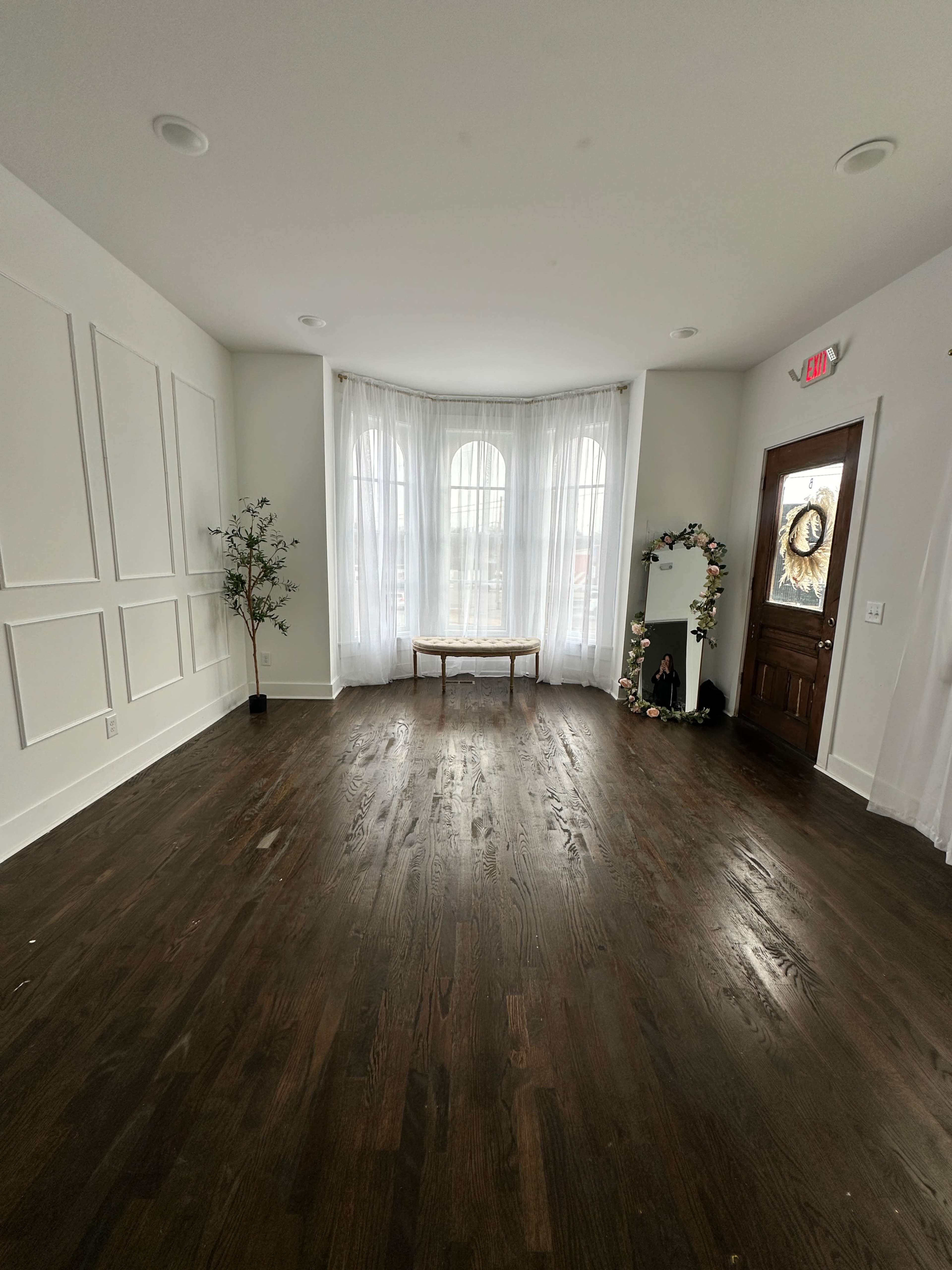 The image shows a bright, empty room with large windows, wooden flooring, a small table in the center, and a decorative mirror against the wall.