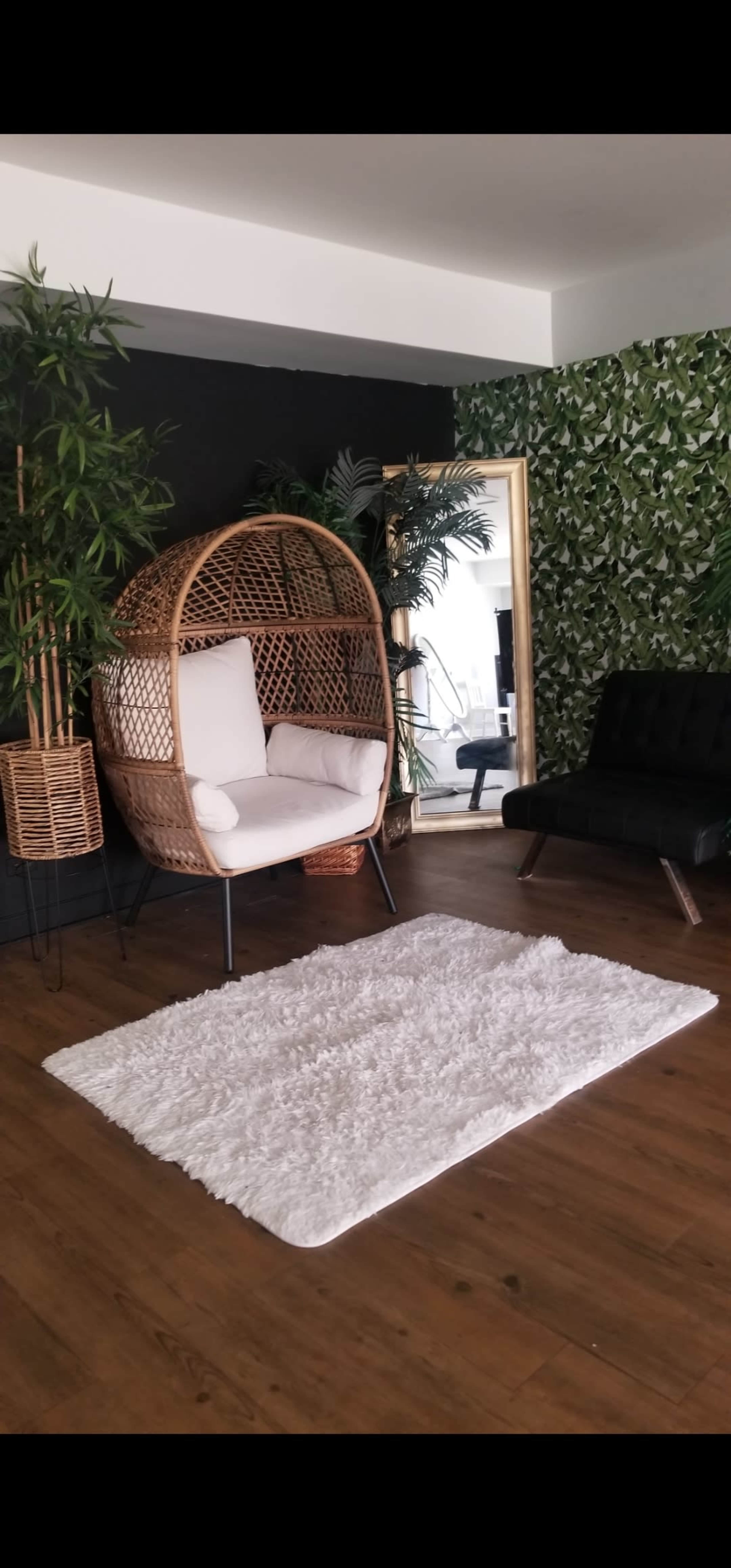 The scene features a stylish seating area with a large wicker chair, a black sofa, a mirror, and a white rug, surrounded by greenery on the walls and in planters.
