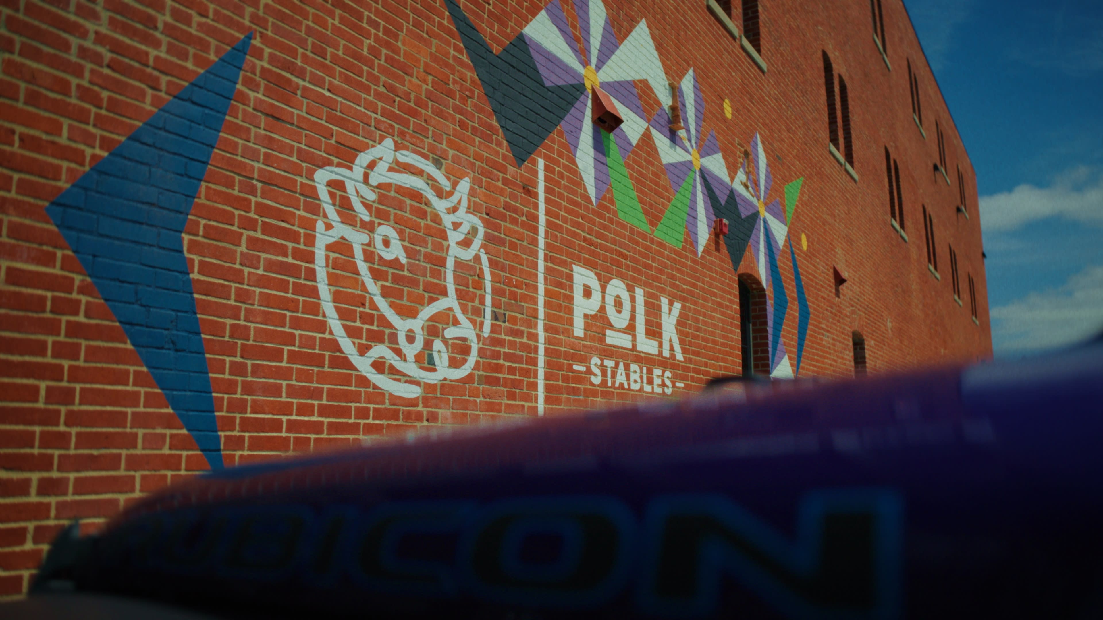 The image features a brick wall displaying a white cow illustration and colorful geometric patterns, alongside the text "POLK STABLES."