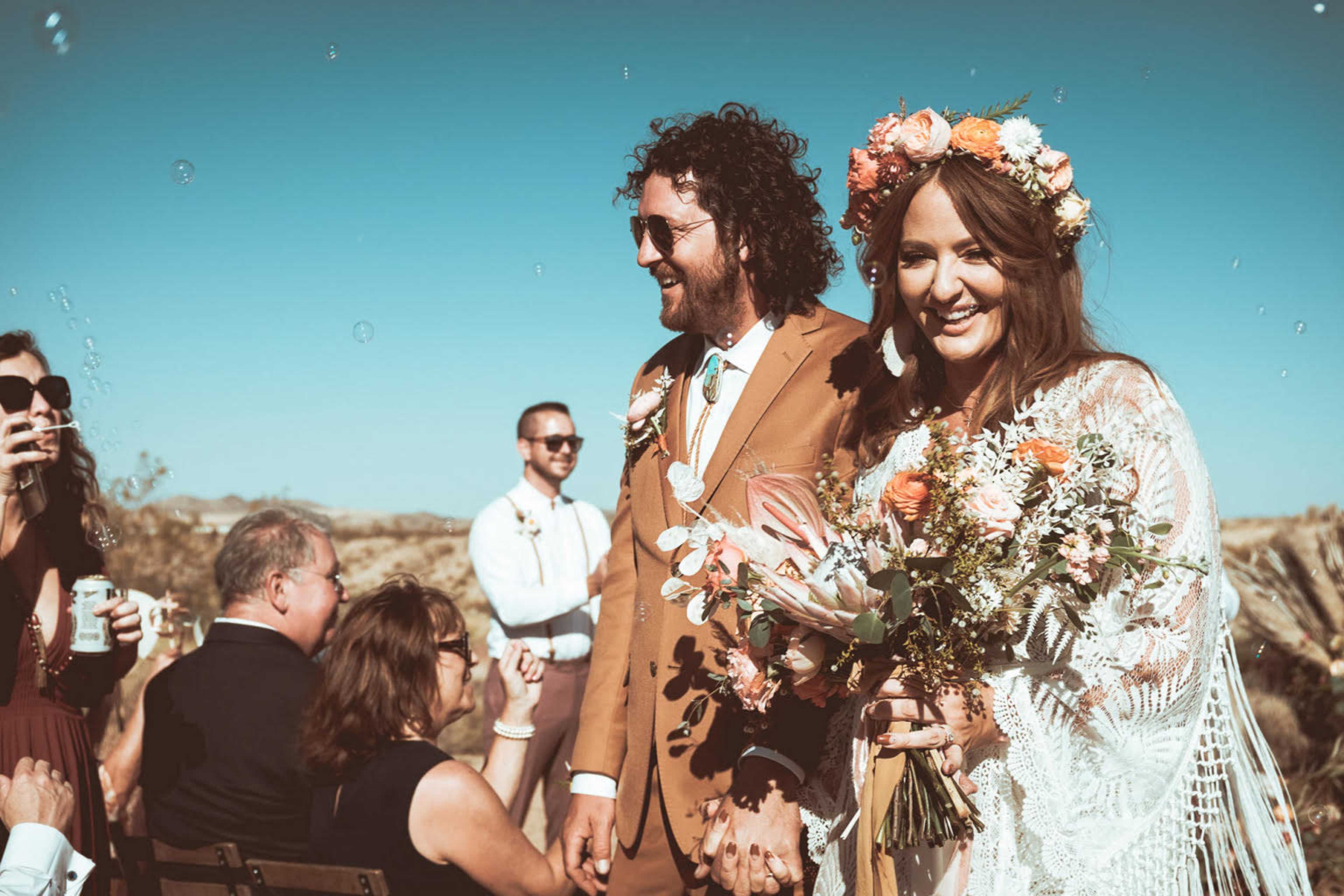 A bride and groom stand together at an outdoor wedding ceremony, surrounded by guests and bubbles, with the bride holding a bouquet of flowers.