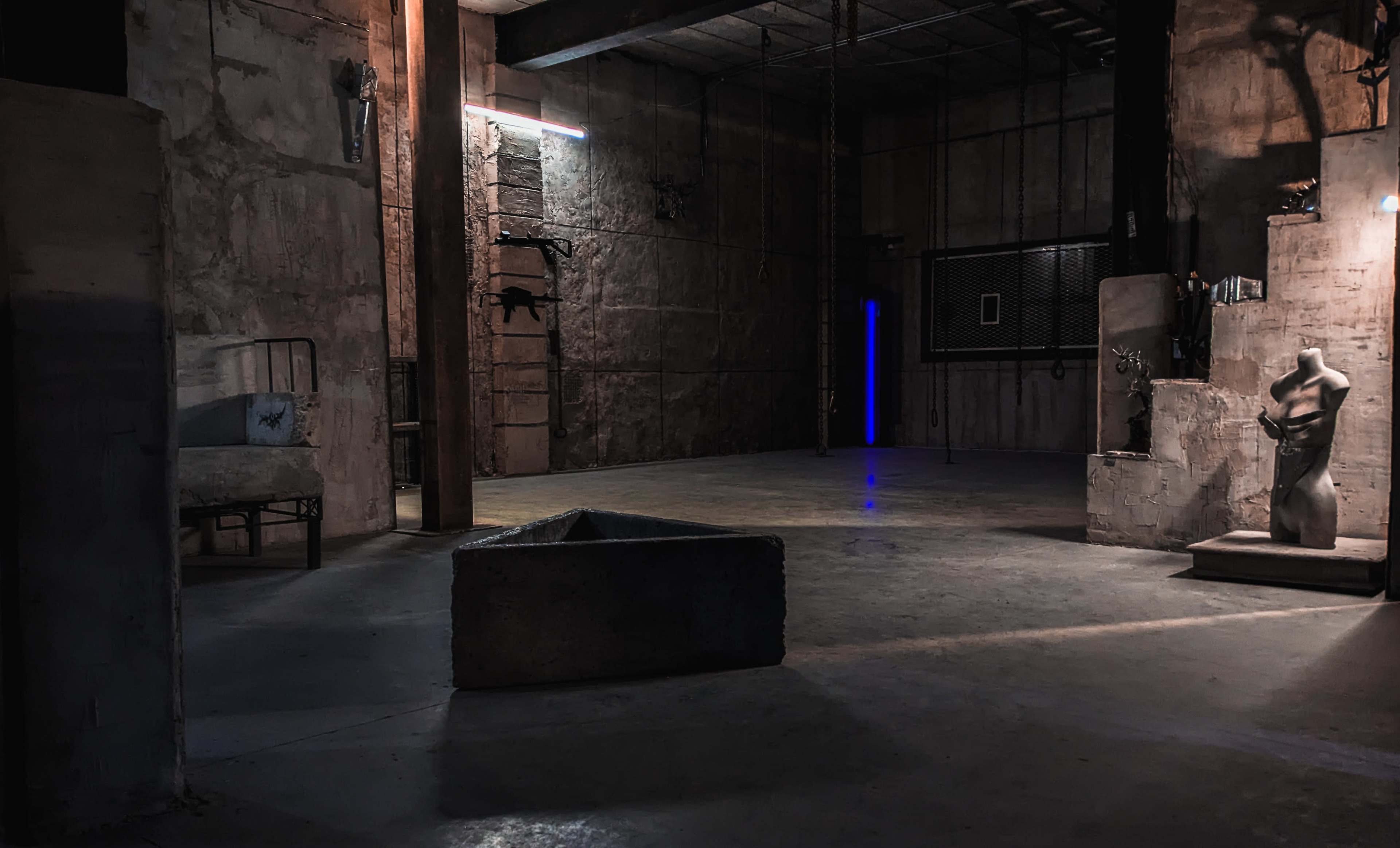 The image shows a dimly lit industrial space with rough concrete walls, a dark sculpture in the center, and a blue light illuminating a corner.