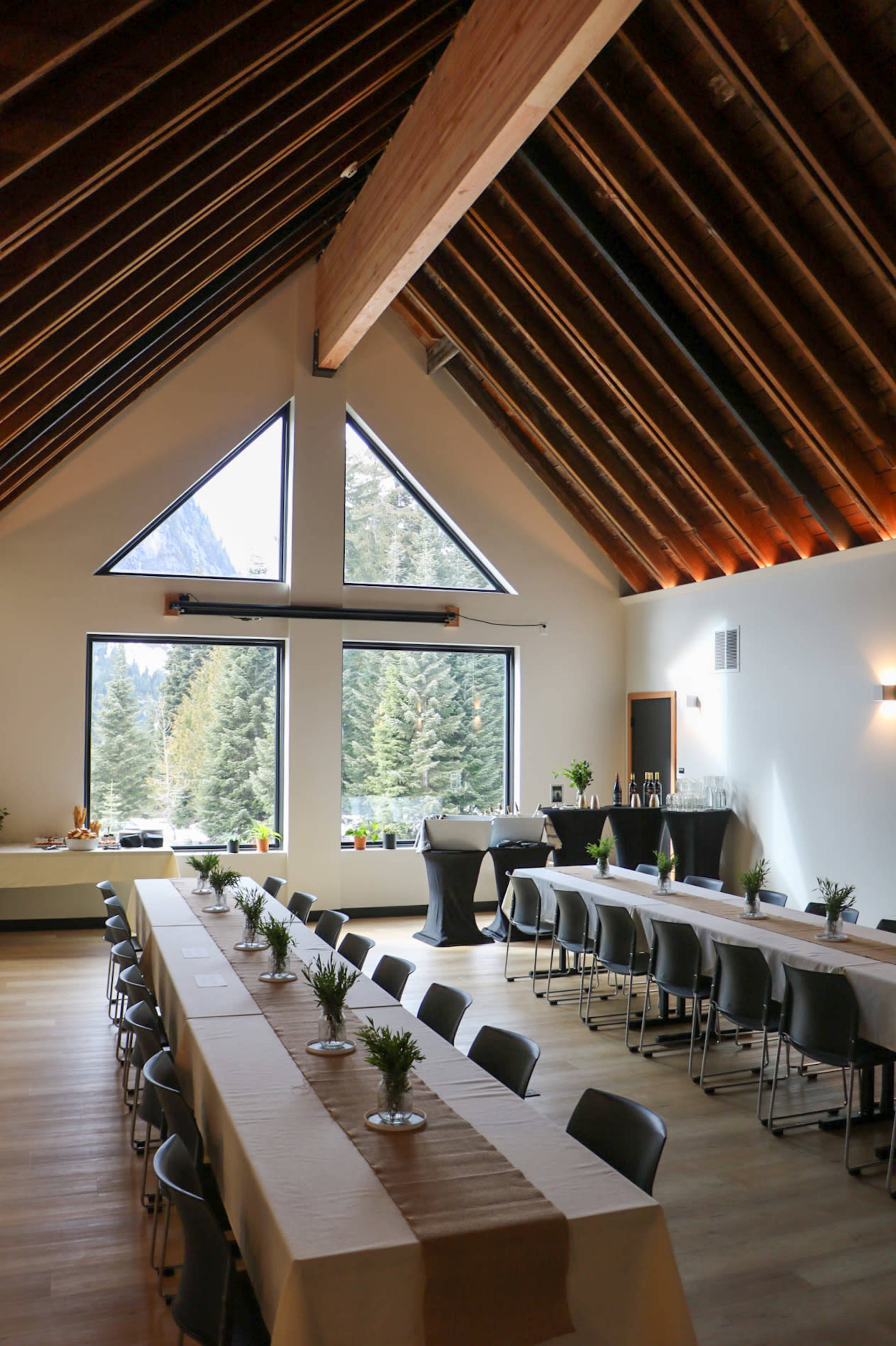 The image shows a spacious indoor event space with large windows showcasing a view of trees, featuring two long tables set up for a gathering.