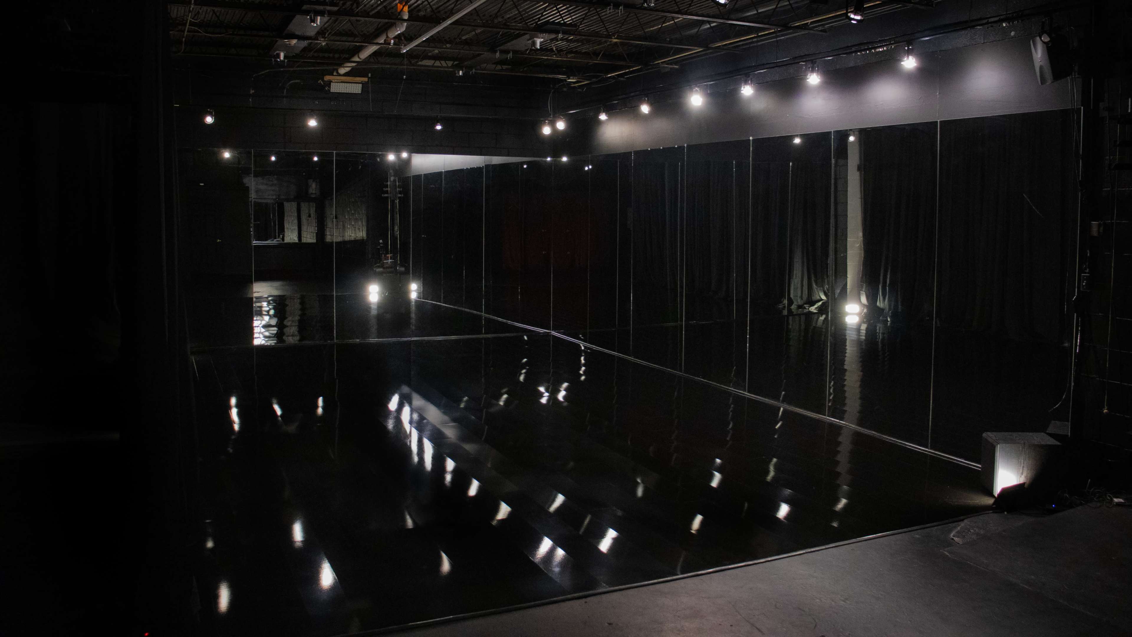 The image shows a dimly lit dance studio with large mirrors lining the walls and spotlights reflecting on the polished floor.