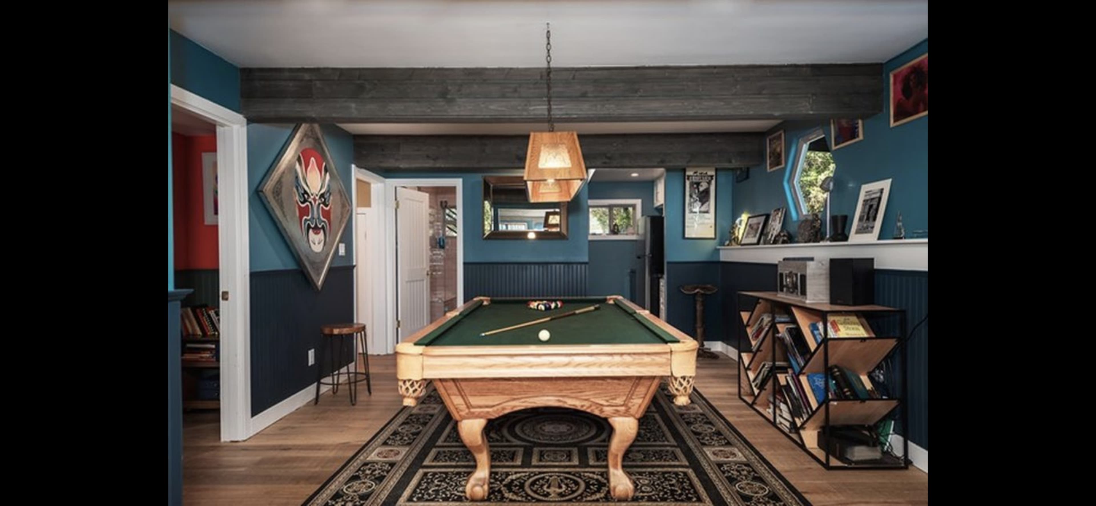 The image shows a billiards room featuring a wooden pool table centered on a patterned rug, with blue walls adorned with artwork and a hanging light fixture above.