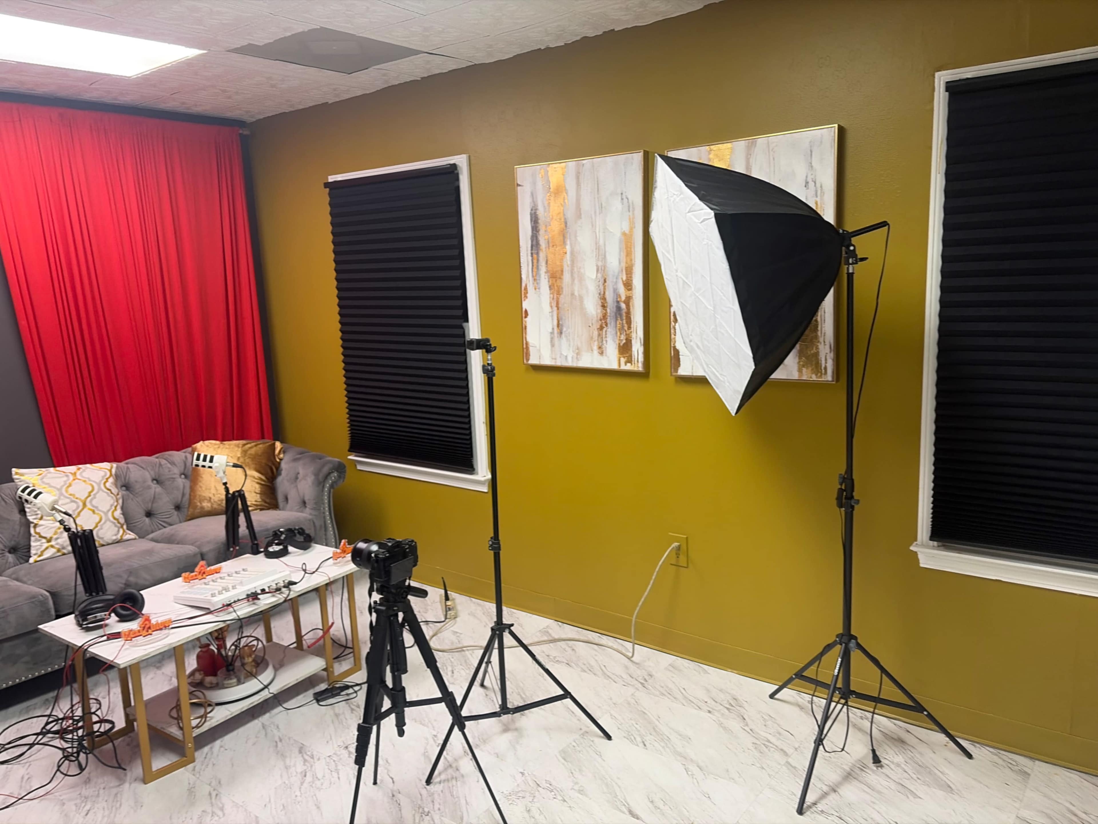 The image shows a room set up for video recording, featuring a gray couch, two windows with black blinds, a tripod with a camera, and softbox lighting.