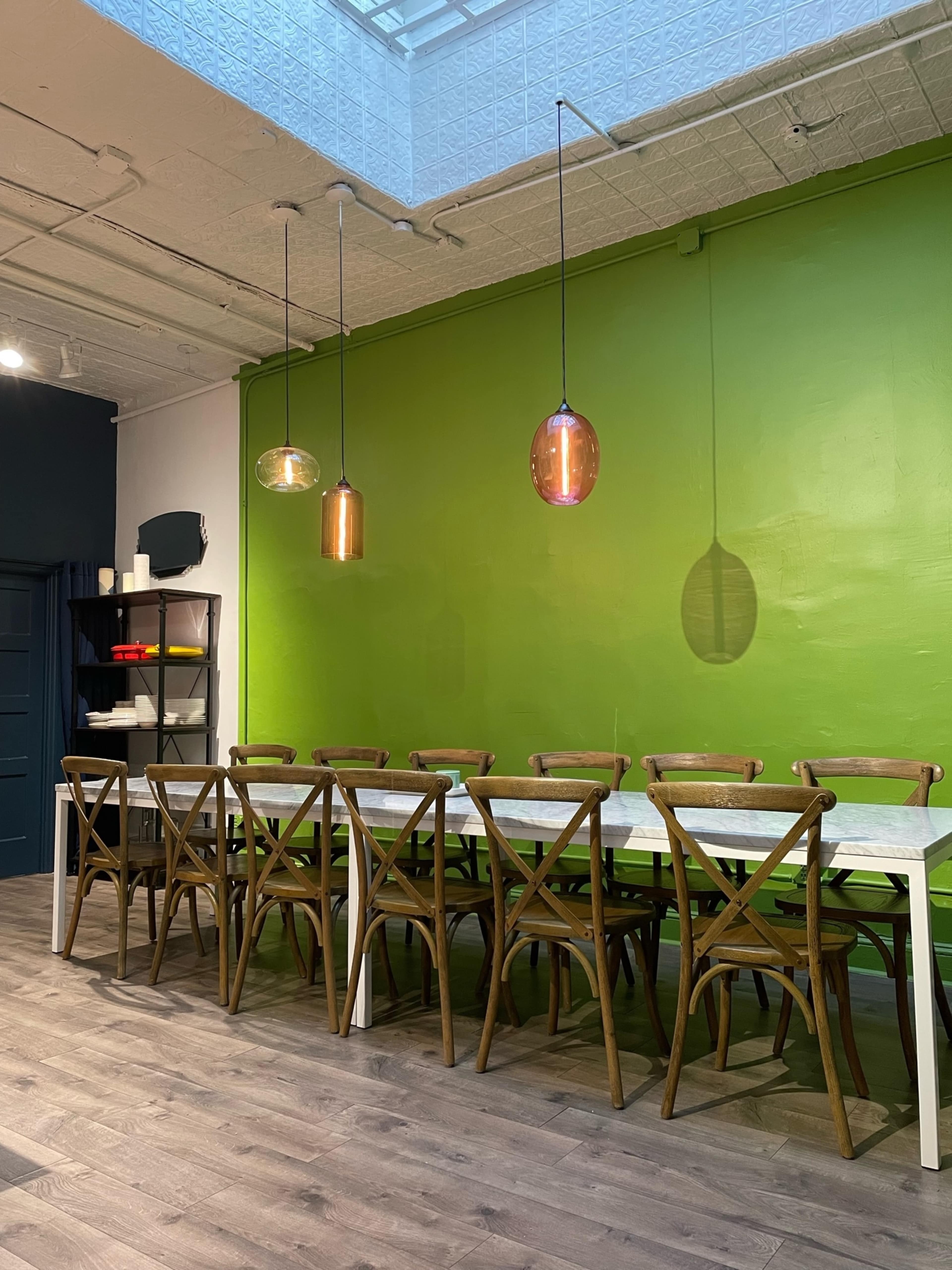 A long, white table with wooden chairs arranged along a bright green wall, illuminated by copper pendant lights.