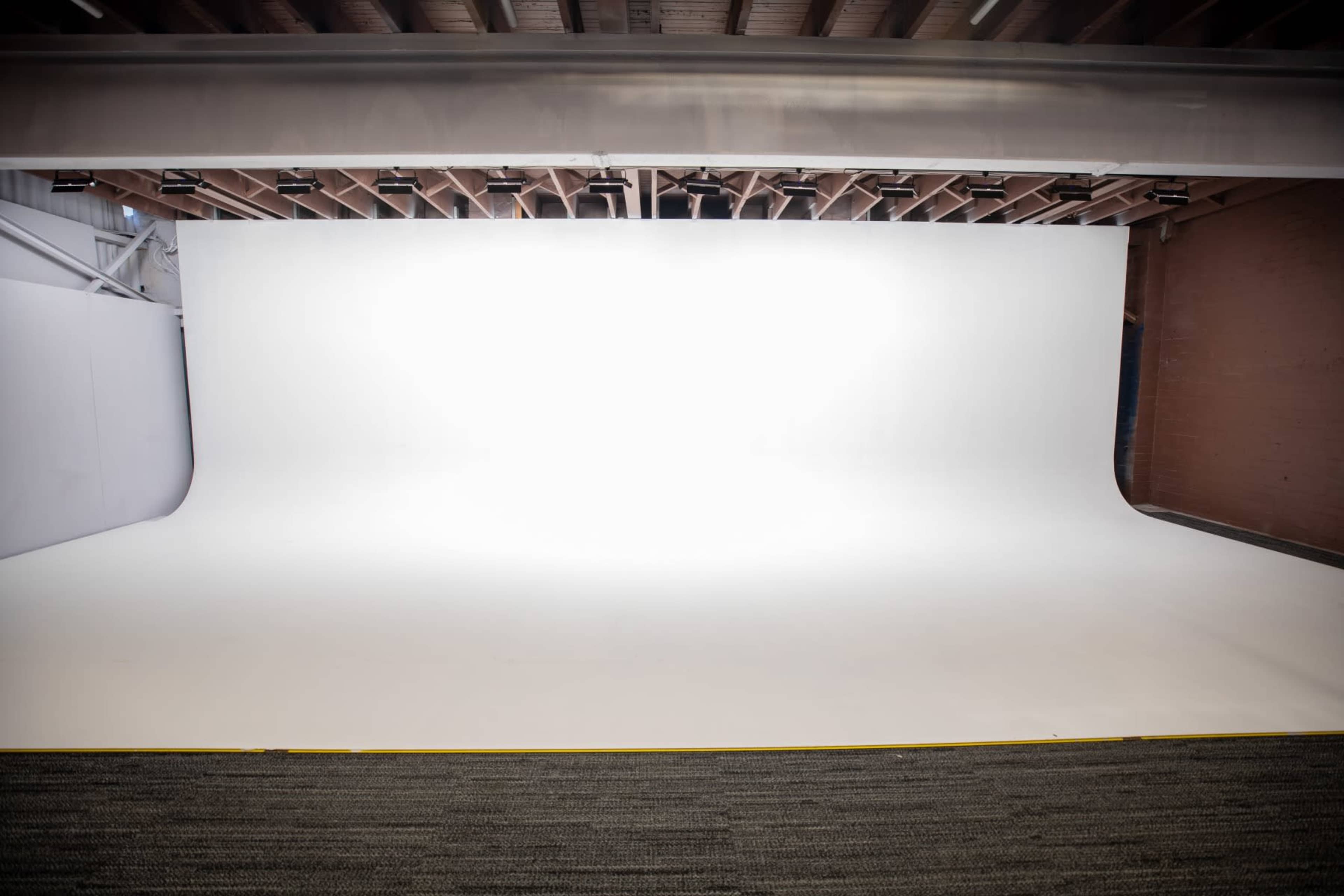 The image shows a spacious photography studio featuring a large, seamless white backdrop and a soft carpeted floor.