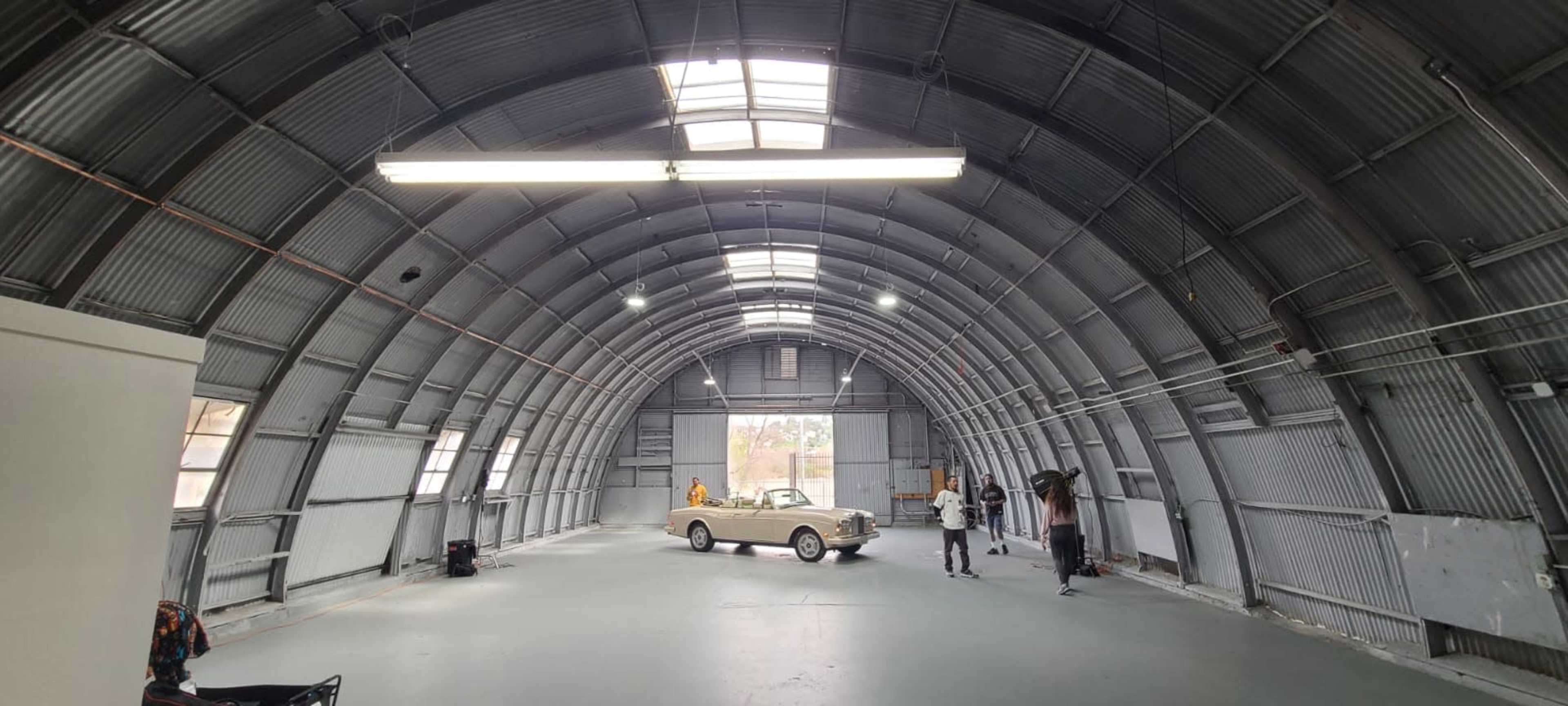 A large, arched metal building houses a few people and a car on a gray floor.