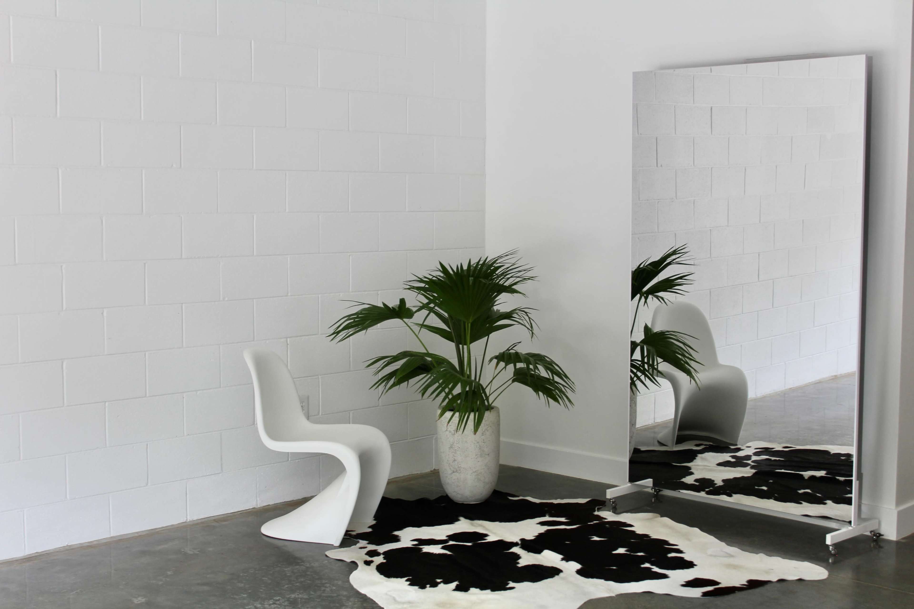 The image shows a minimalist room with a white wall, featuring a curved white chair, a large mirror, a plant in a pot, and a black-and-white cowhide rug on the floor.