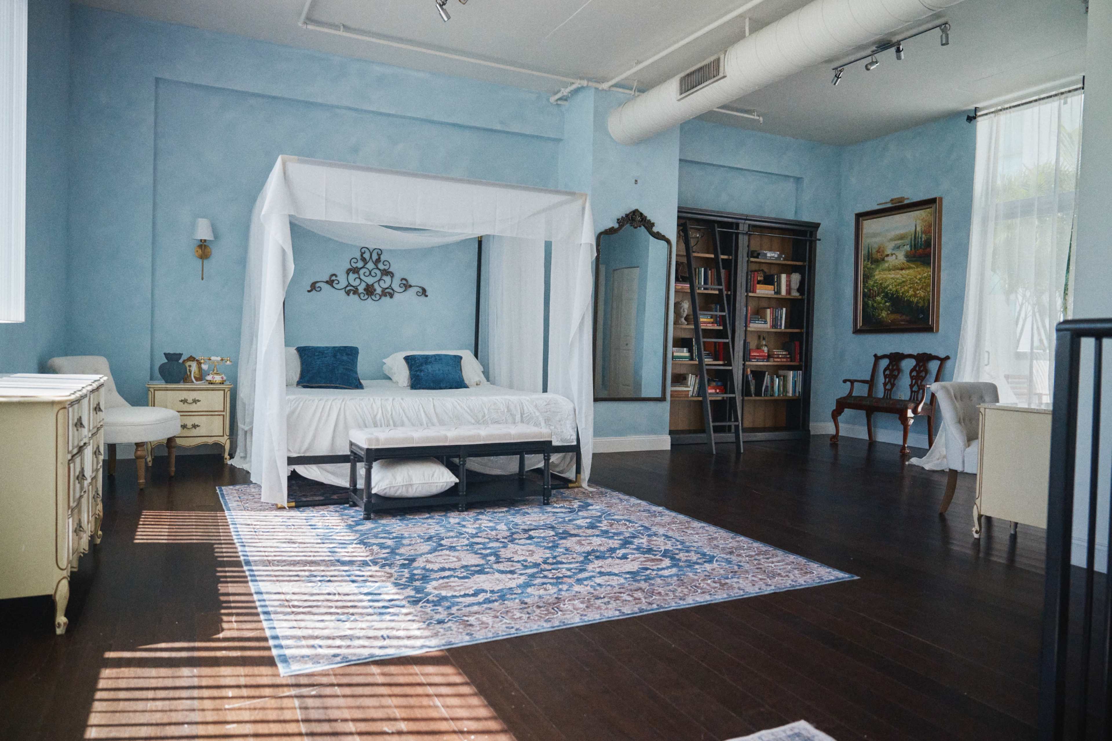 The image shows a light blue bedroom featuring a four-poster bed, a bookcase filled with books, and a large painting on the wall.