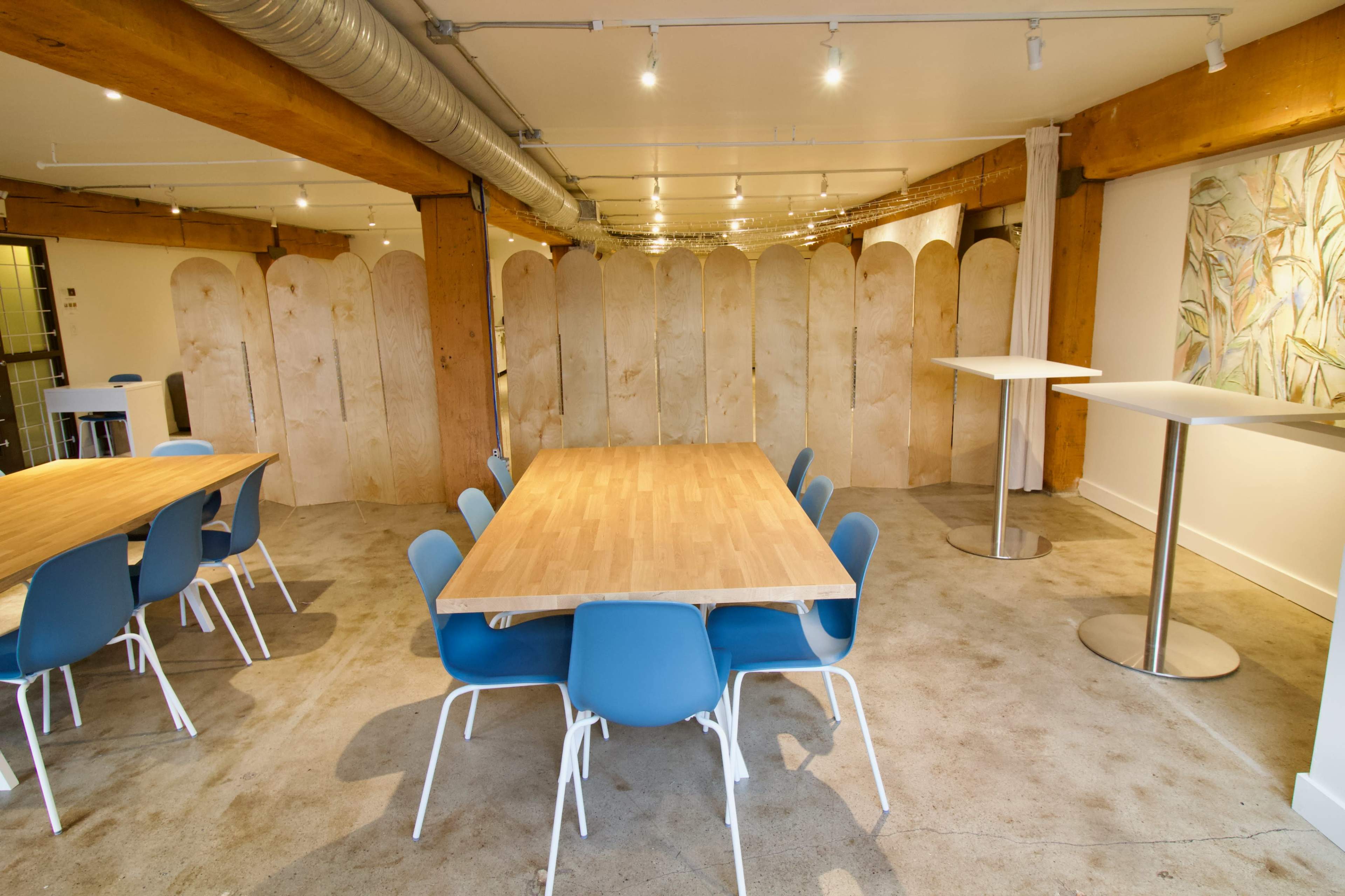 The image shows a modern, spacious room with wooden tables and blue chairs, separated by wooden partitions.