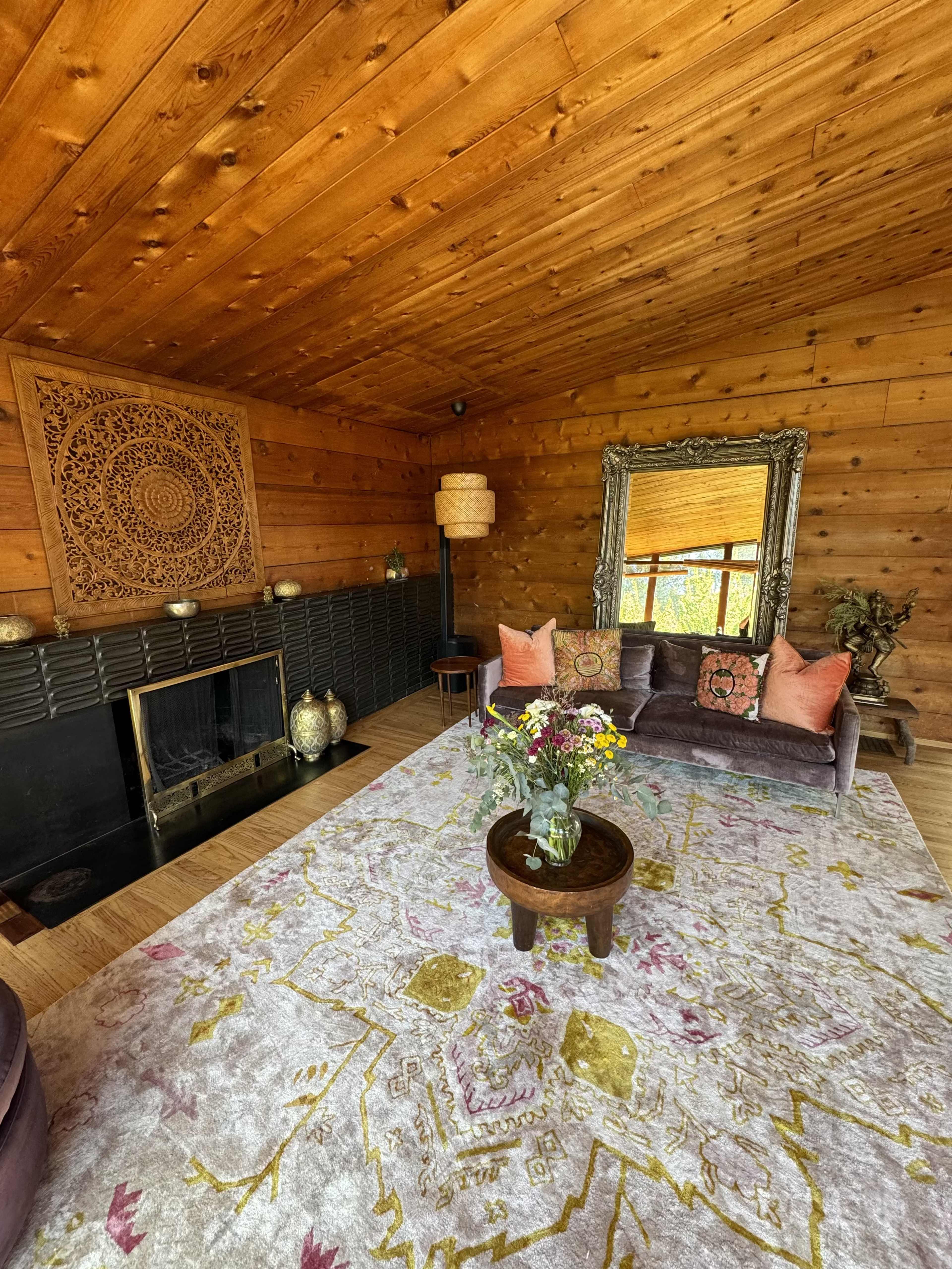 A cozy living room with wooden walls, a fireplace, a large mirror, and a floral area rug.