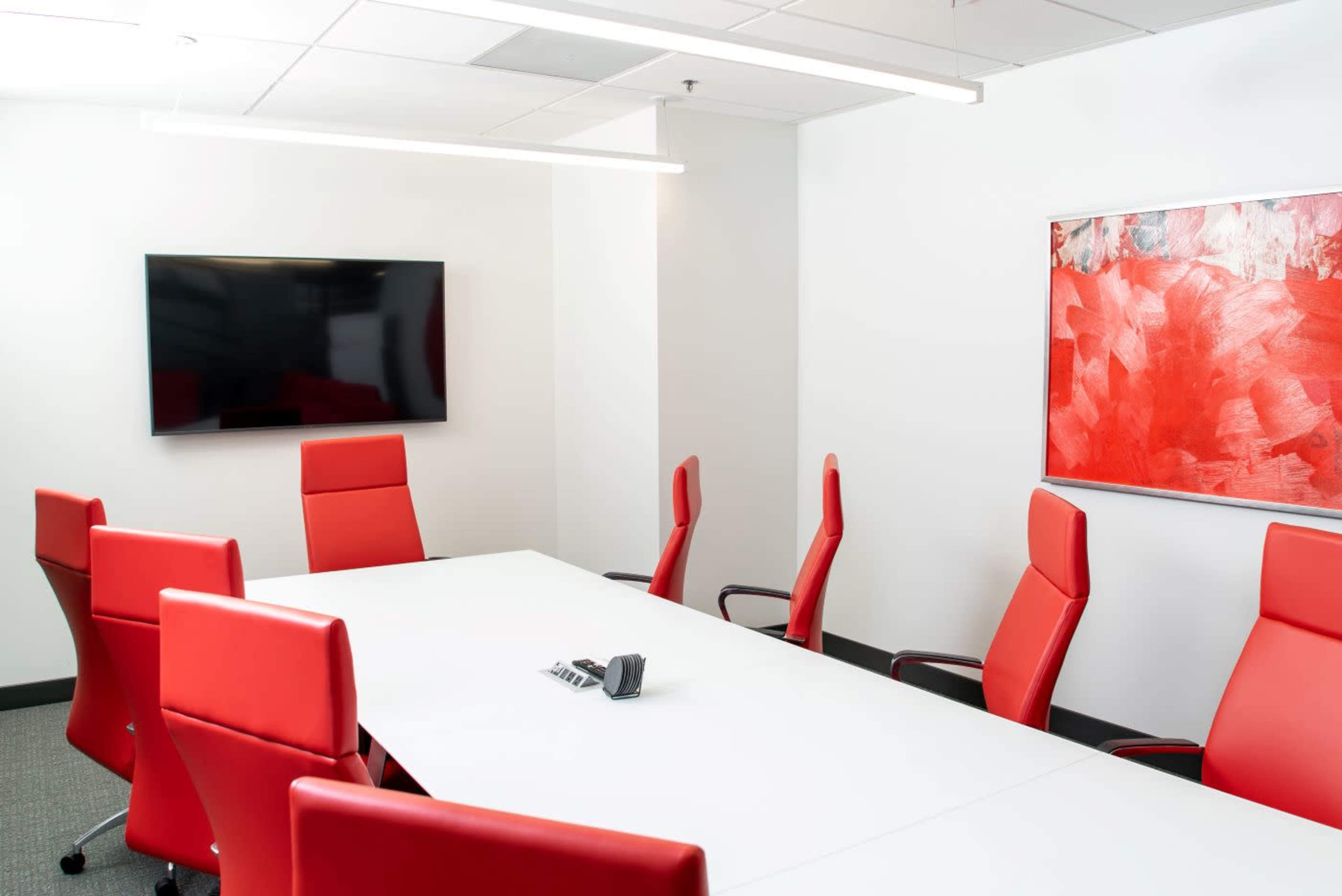 A modern conference room features a rectangular white table surrounded by red ergonomic chairs, with a wall-mounted TV and an abstract red artwork.