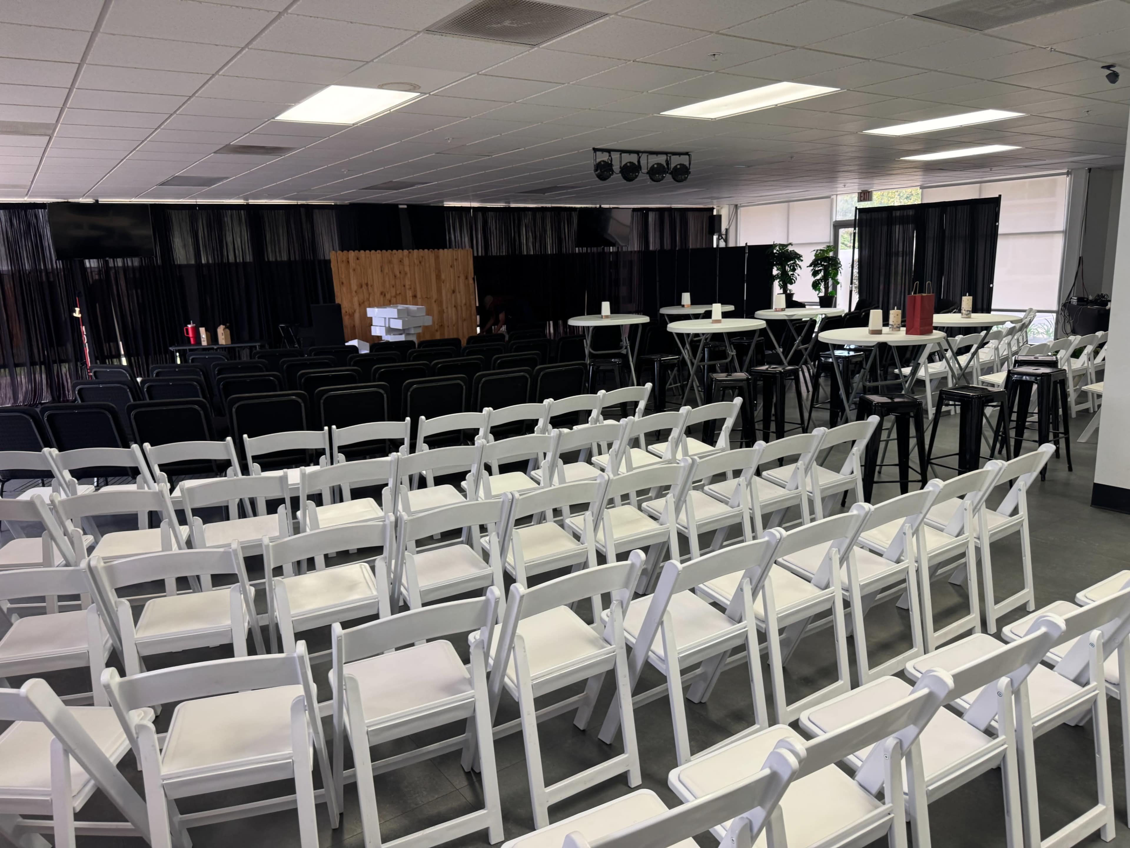 The image shows a room set up for an event with rows of white folding chairs facing a stage area, along with tall tables and bar stools positioned nearby.
