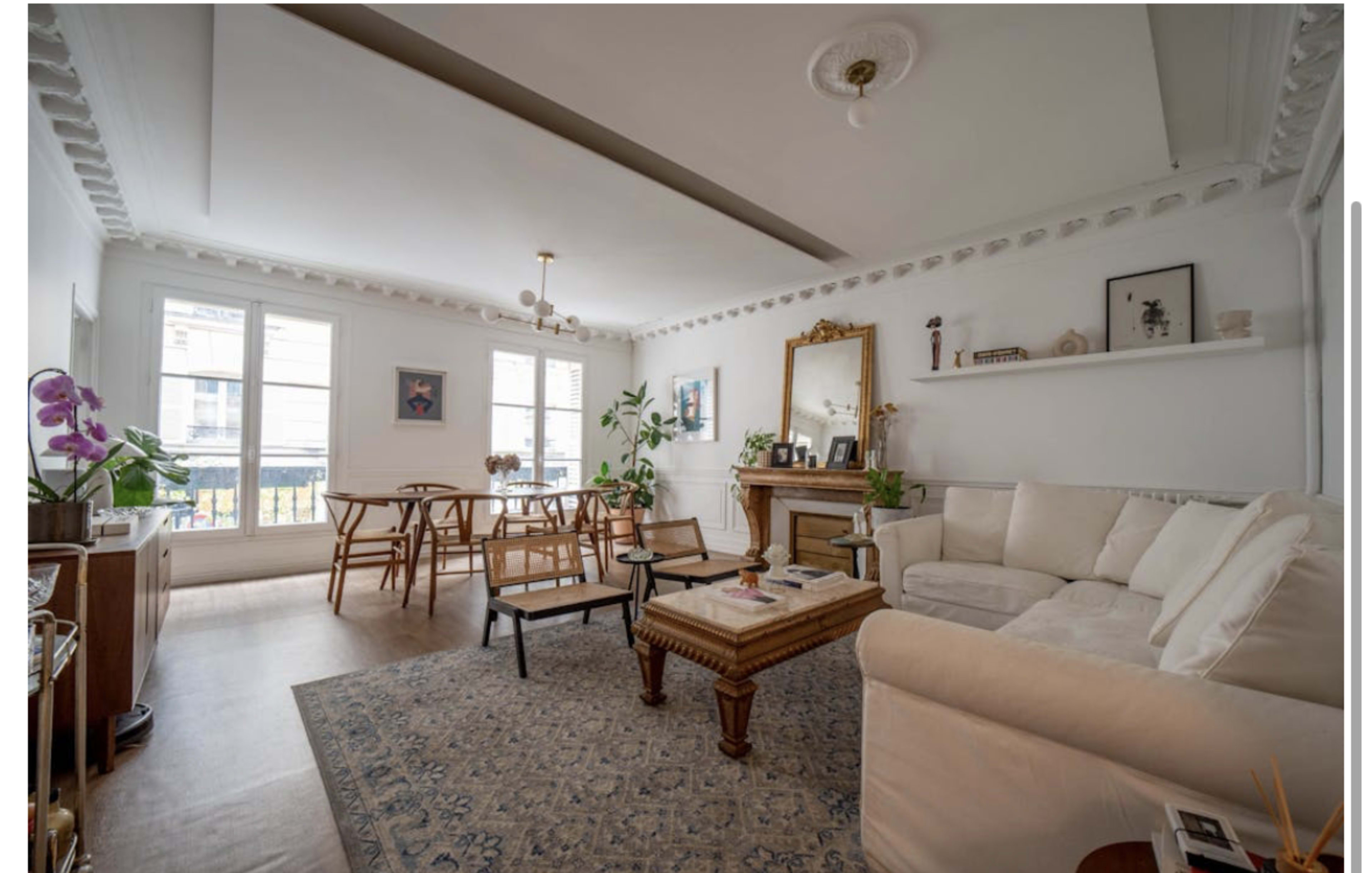 The image shows a bright living room with white walls, a large window, a sectional sofa, wooden furniture, and decorative plants.