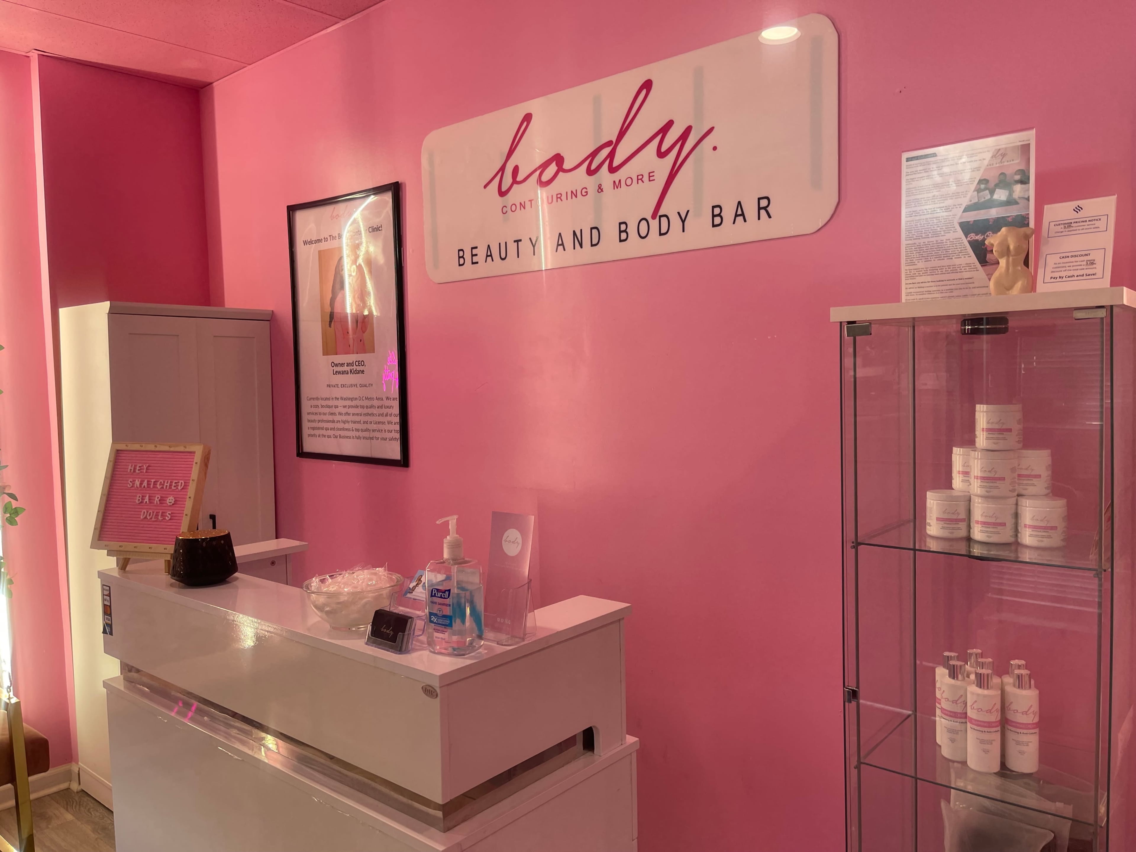 The interior of a beauty and body bar with a pink wall, a reception desk, and display shelves featuring beauty products.