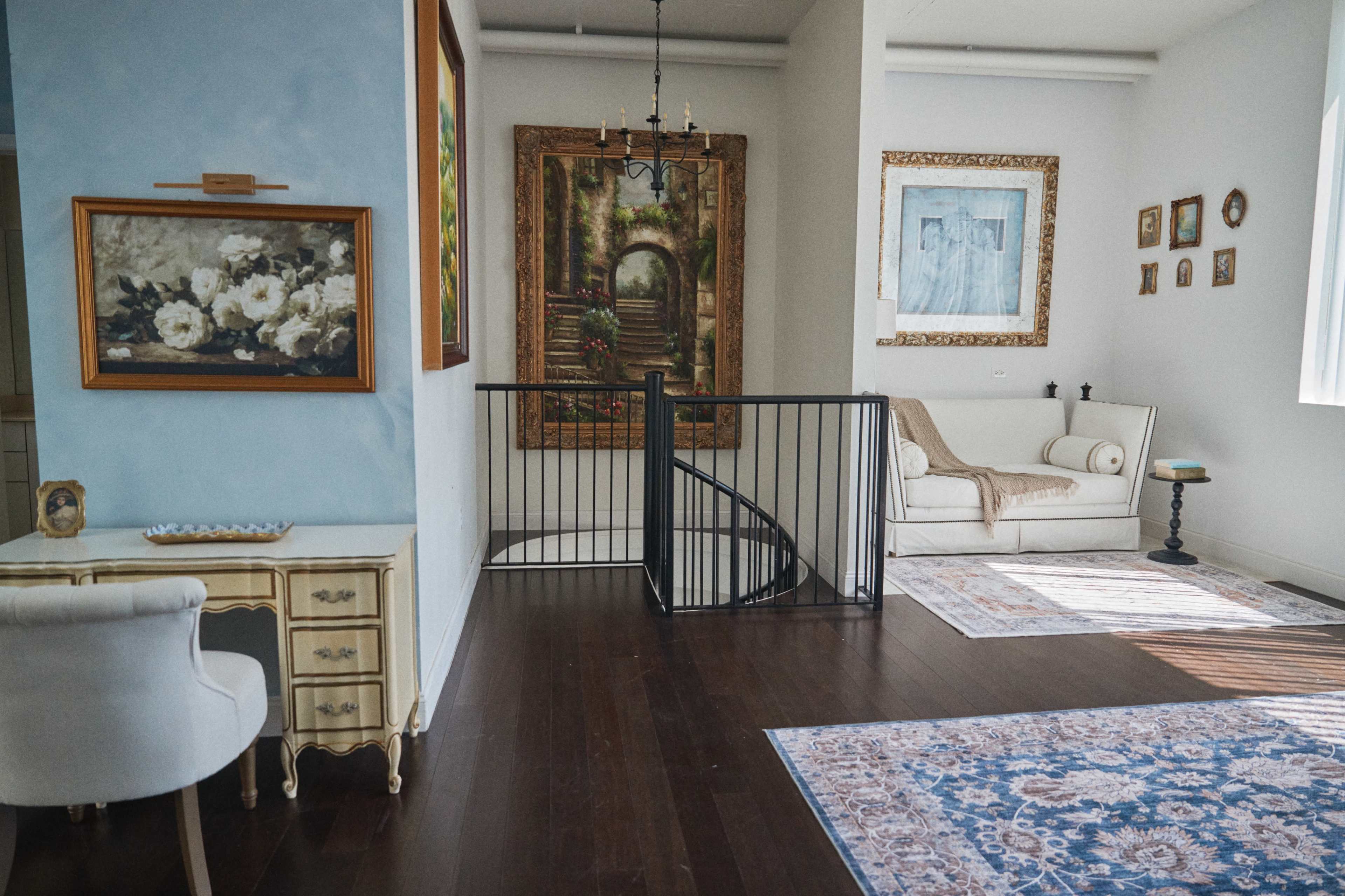 The image shows an interior space featuring a staircase, a cream-colored sofa, various artworks on the walls, a decorative rug, and furniture arranged around the room.