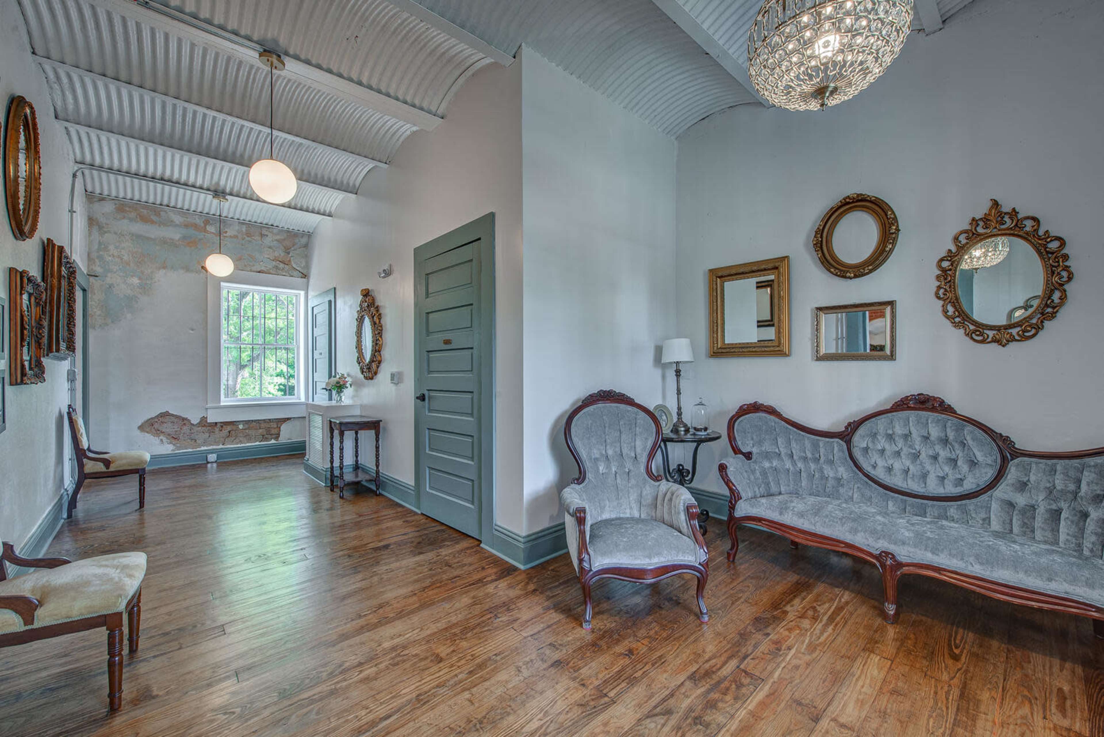 A spacious hallway with a light-colored wall, wood flooring, a vintage-style seating area, and several decorative mirrors.