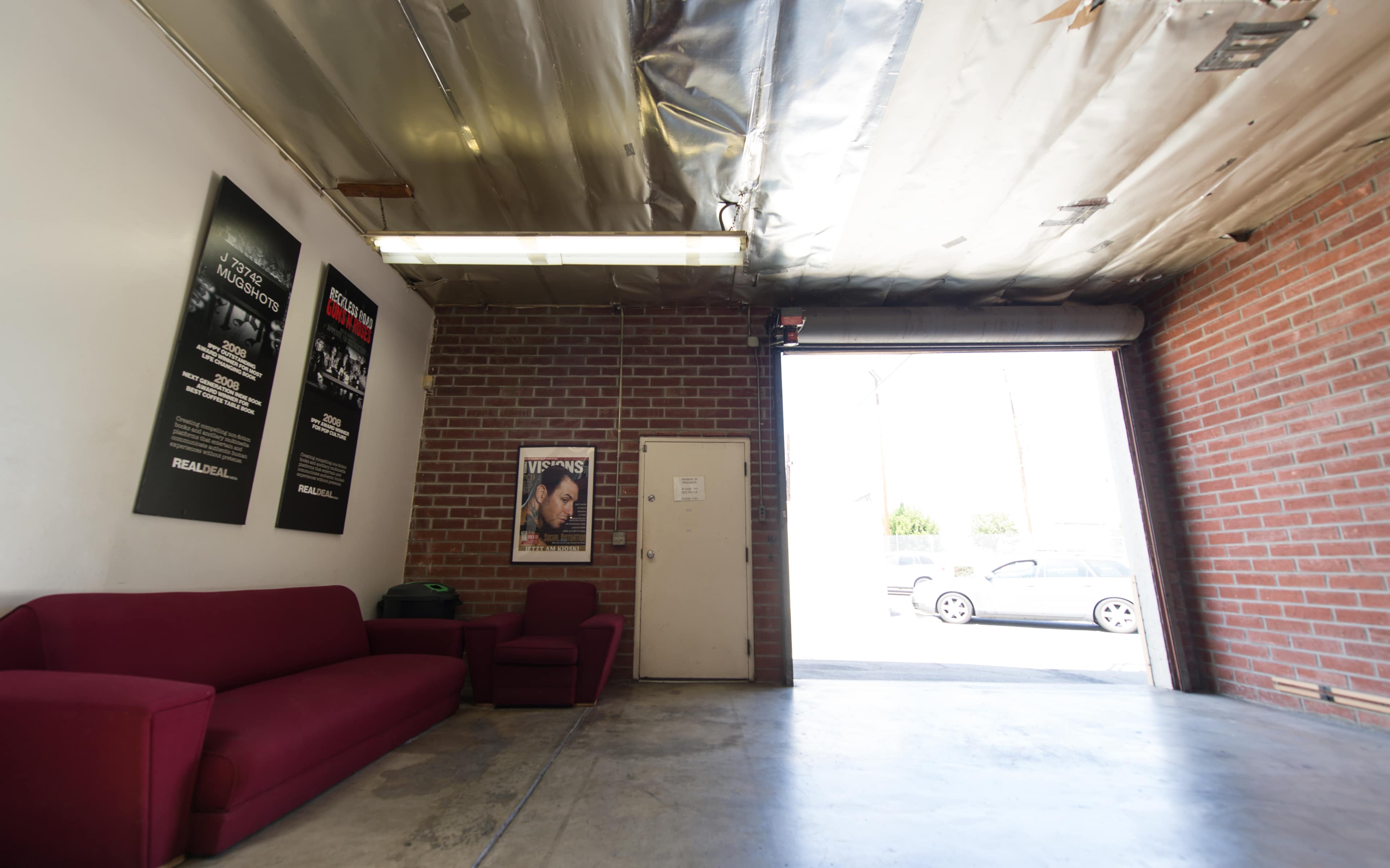 The image shows an interior of a garage-like space with a red couch, a chair, a door, and a large opening leading outside.