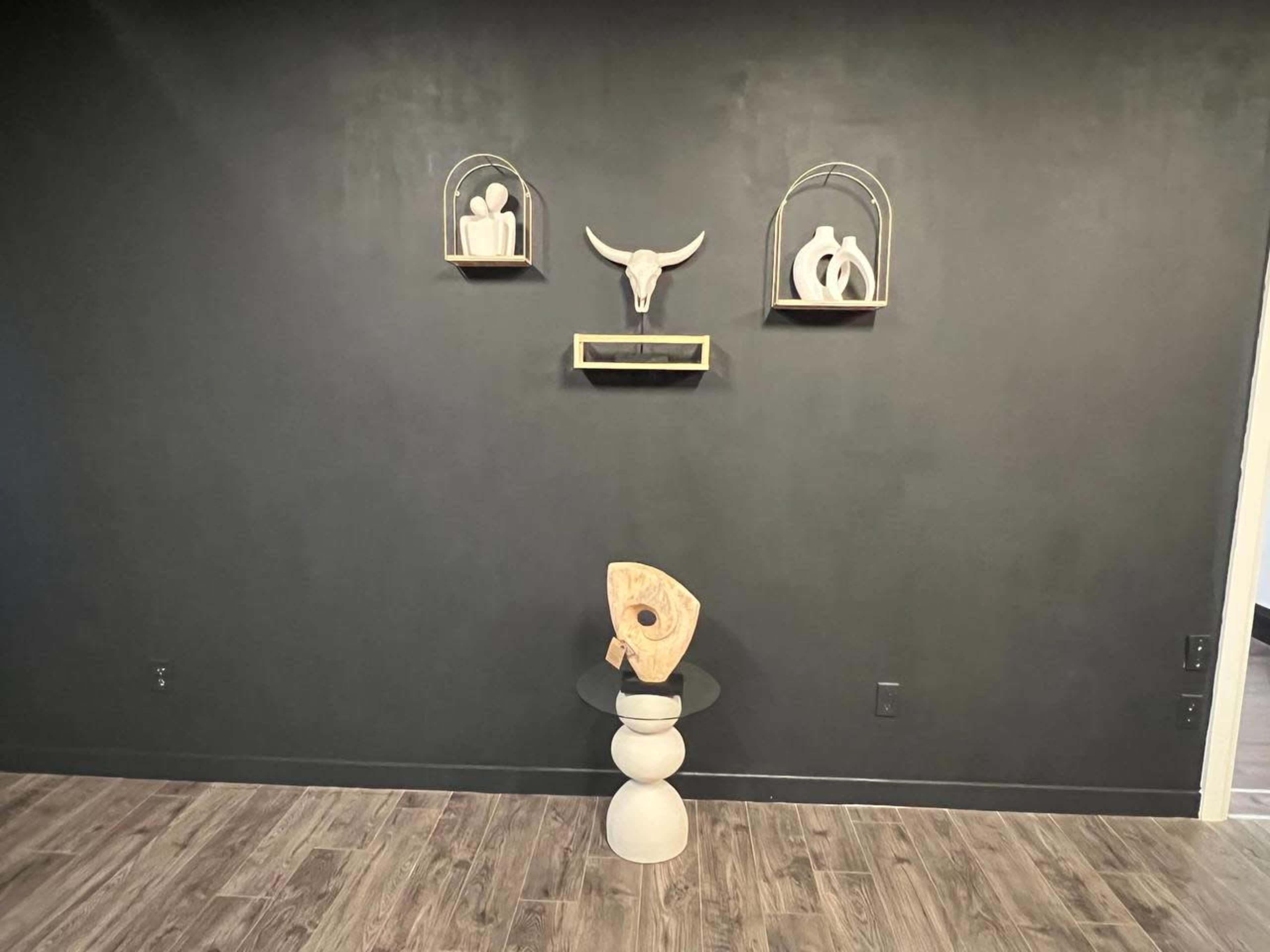 The image shows a dark wall featuring three decorative shelves with minimalist sculptures and a central wooden object on a pedestal.