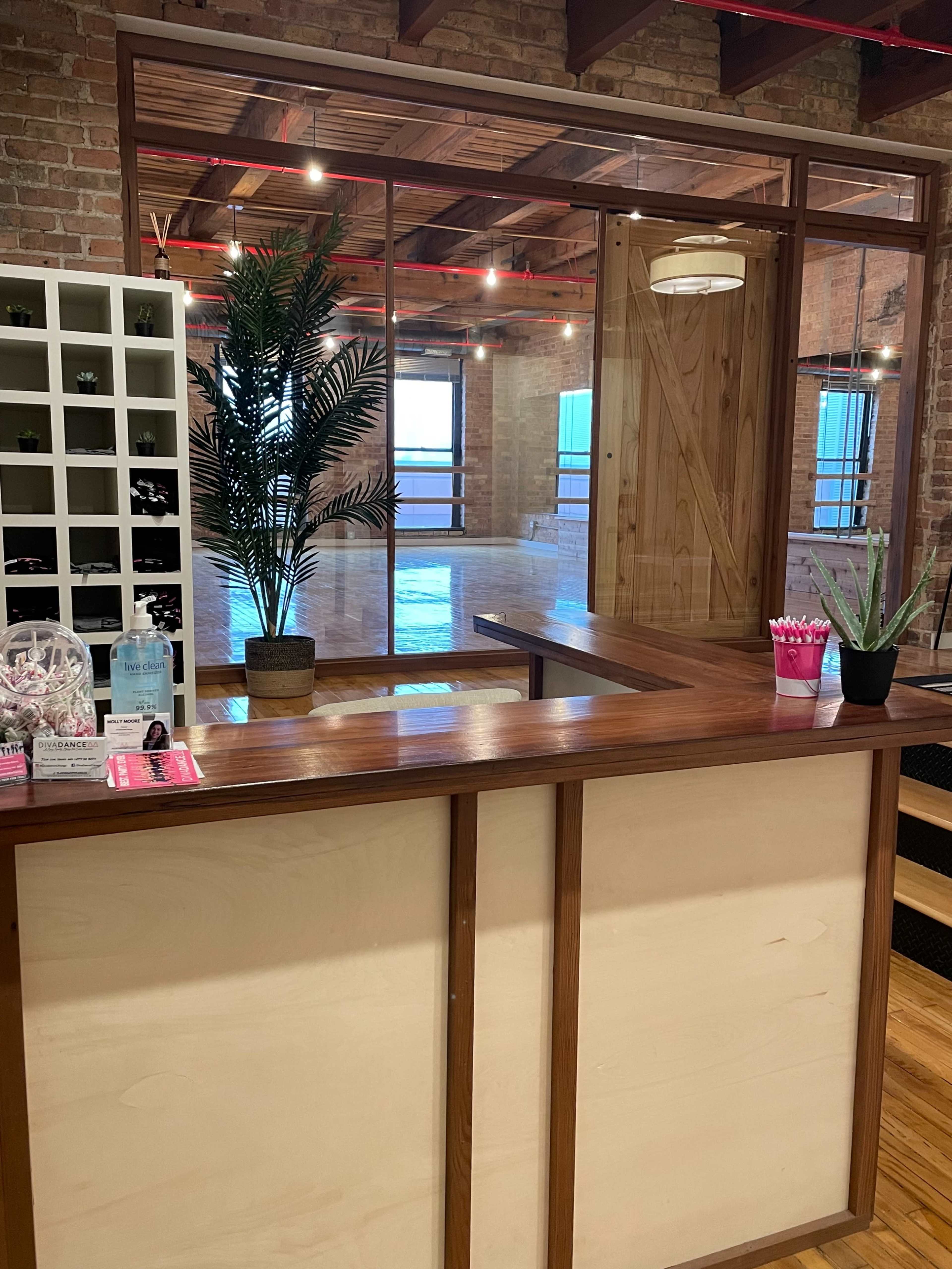 A reception area features a wooden counter with plants and glass panels separating it from a spacious room with wooden flooring and brick walls.