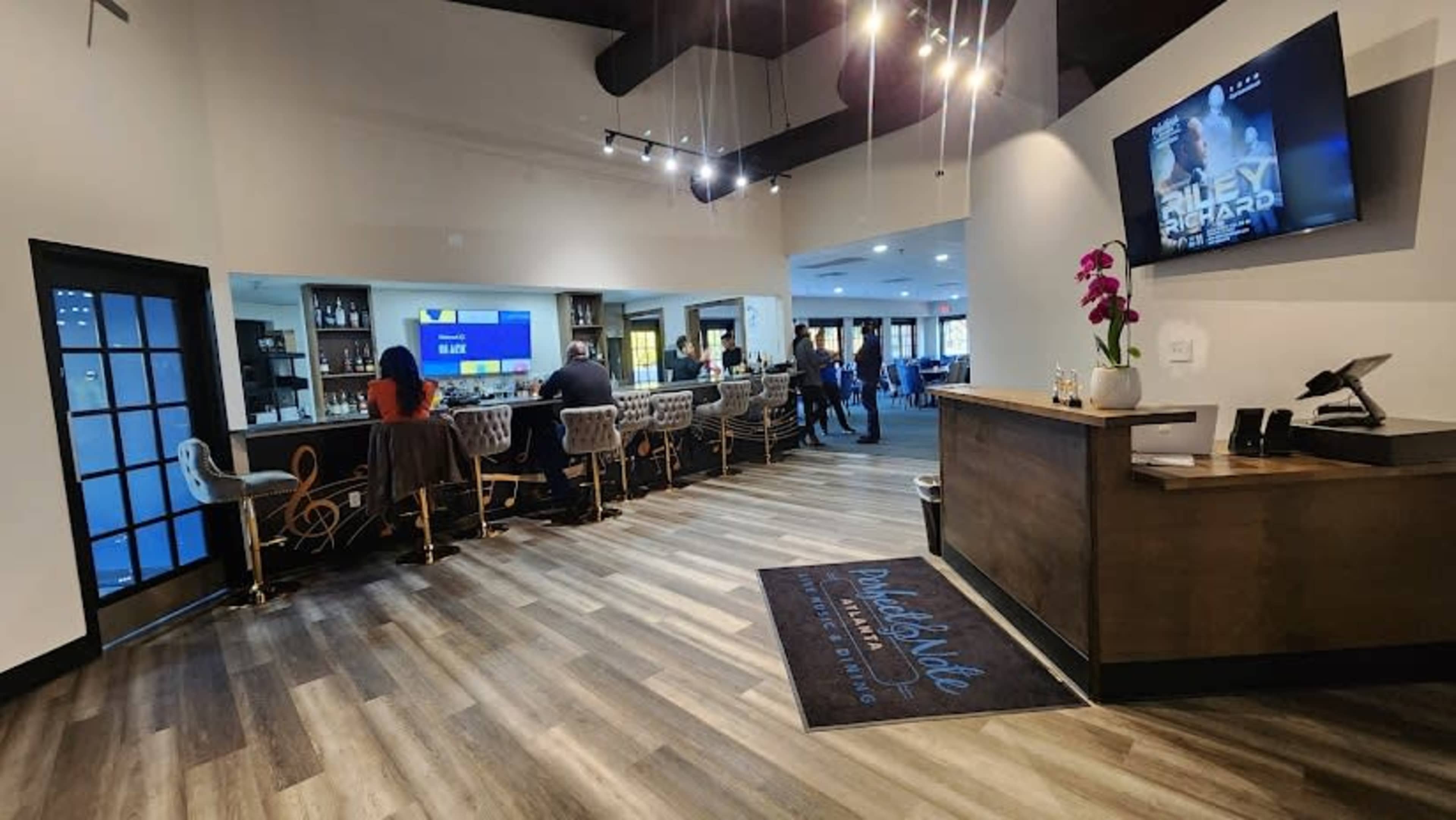 The interior of a modern bar or restaurant with a central bar area and seating, featuring a dark wood reception desk and a flat-screen TV mounted on the wall.