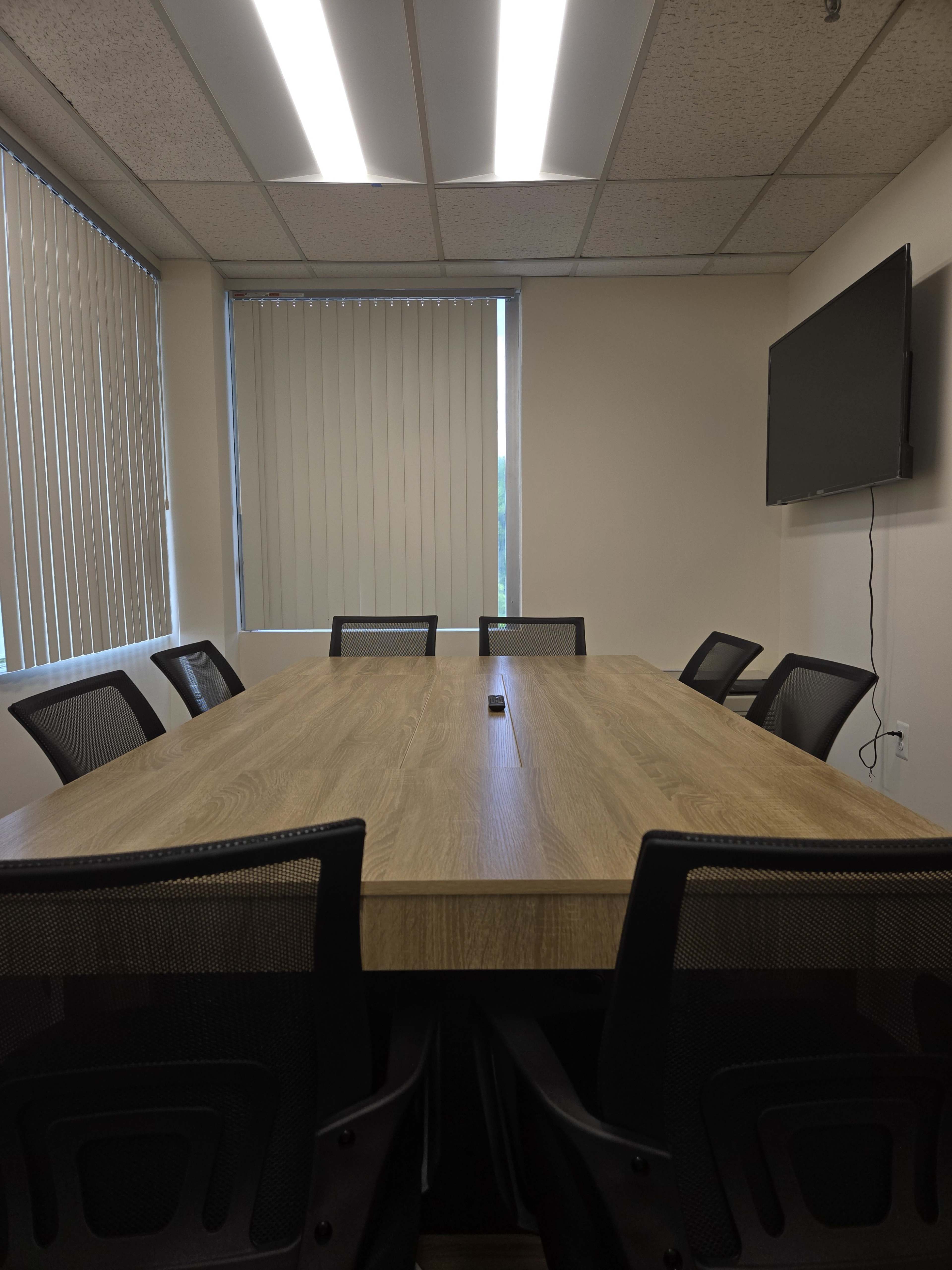 A rectangular wooden conference table is set in a brightly lit meeting room with black mesh chairs arranged around it and a wall-mounted television.