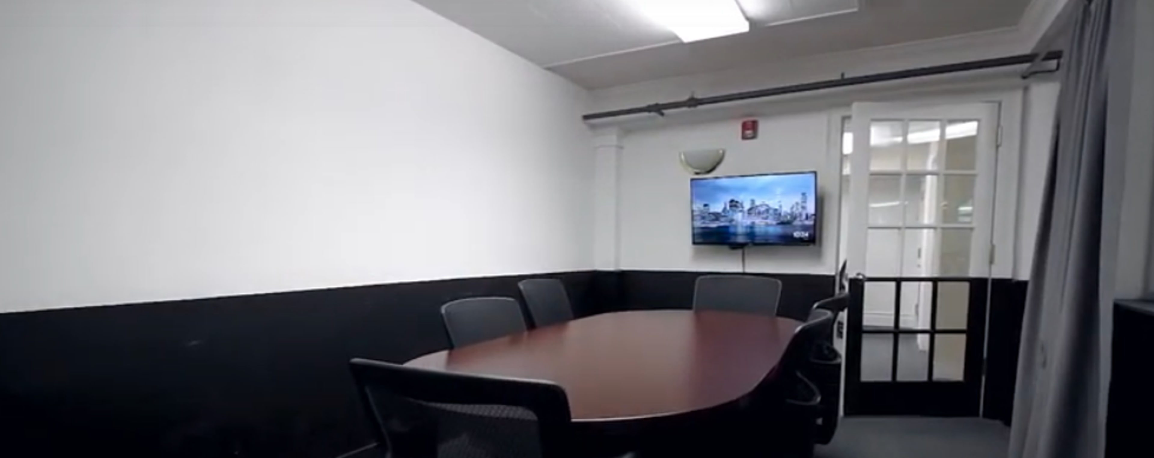 A conference room features a large oval table surrounded by black chairs, with a television mounted on the wall and a door leading to another area.