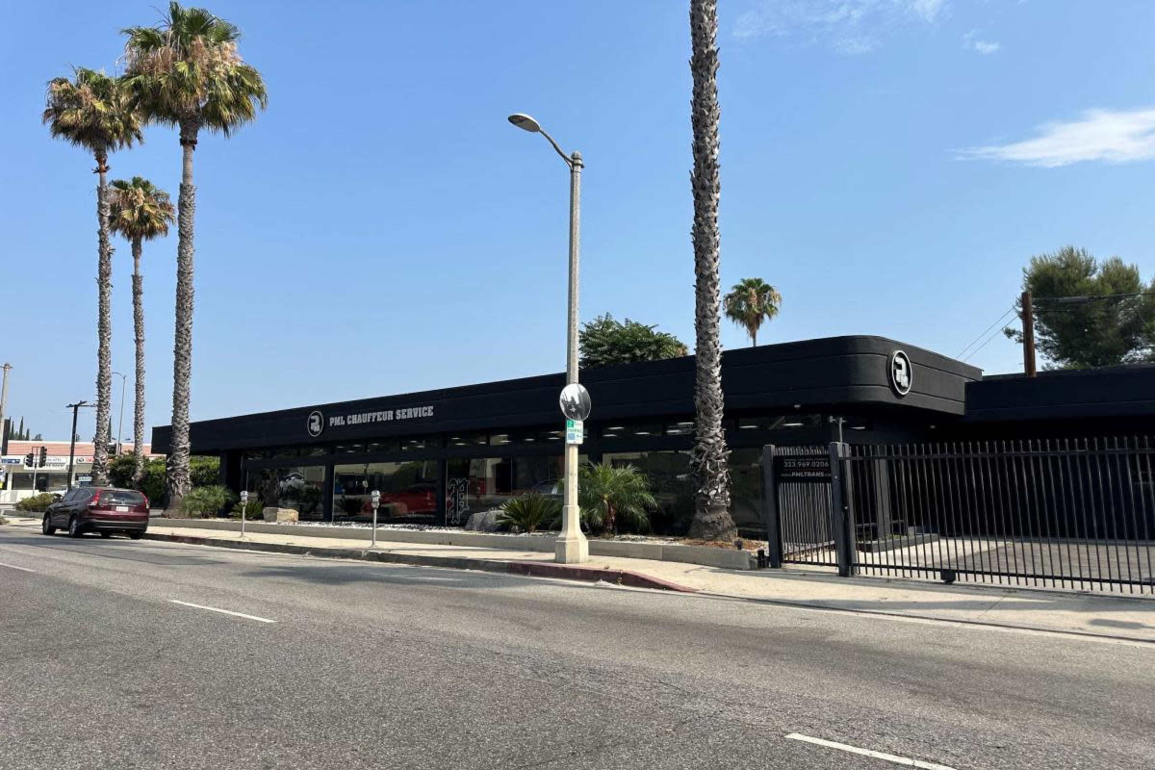 The image shows a modern building with a black exterior, featuring large windows and palm trees in front, located along a road.