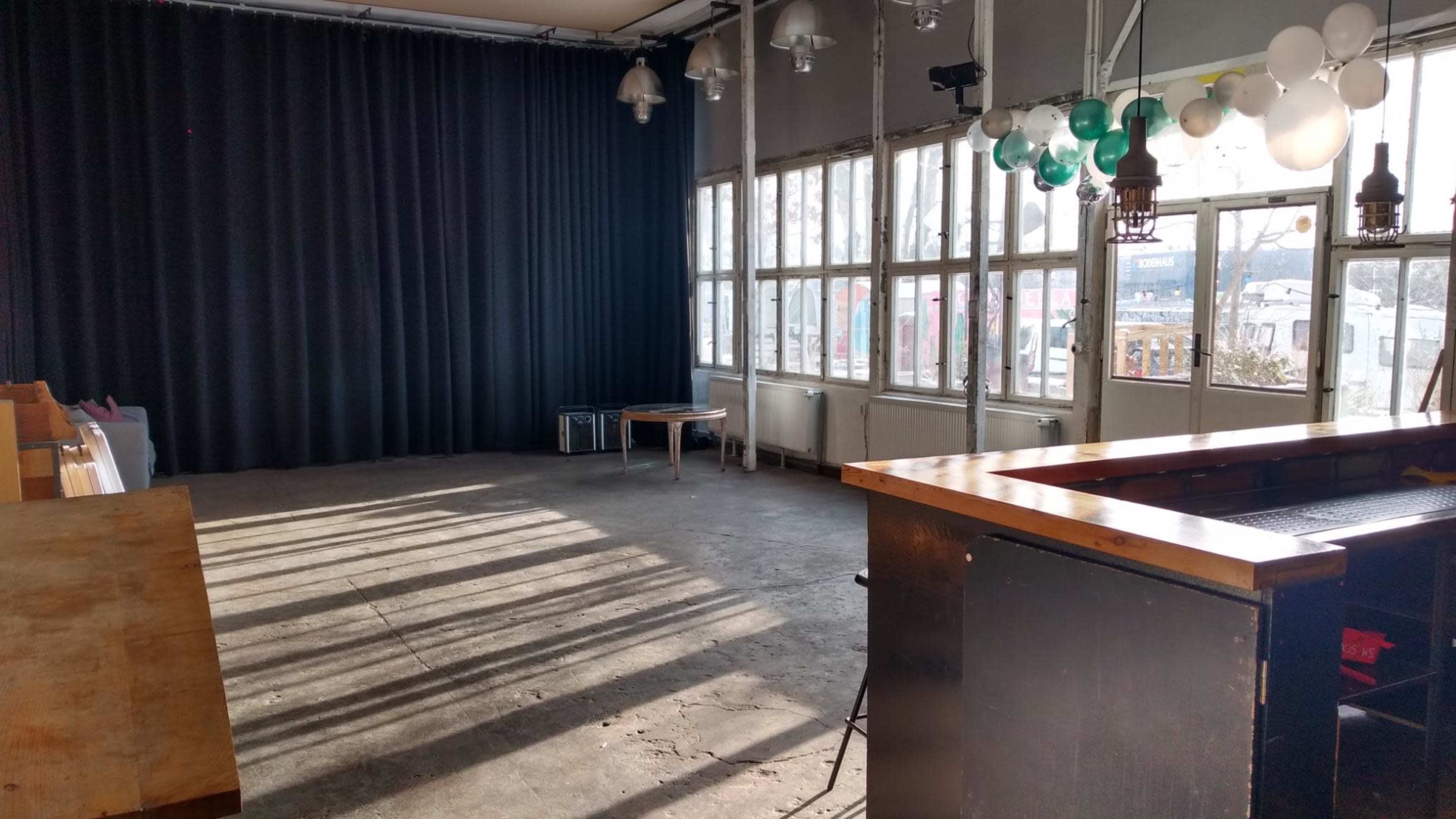 The image shows an empty room with large windows allowing natural light to cast shadows on the concrete floor, featuring a wooden bar and decorations hanging from the ceiling.