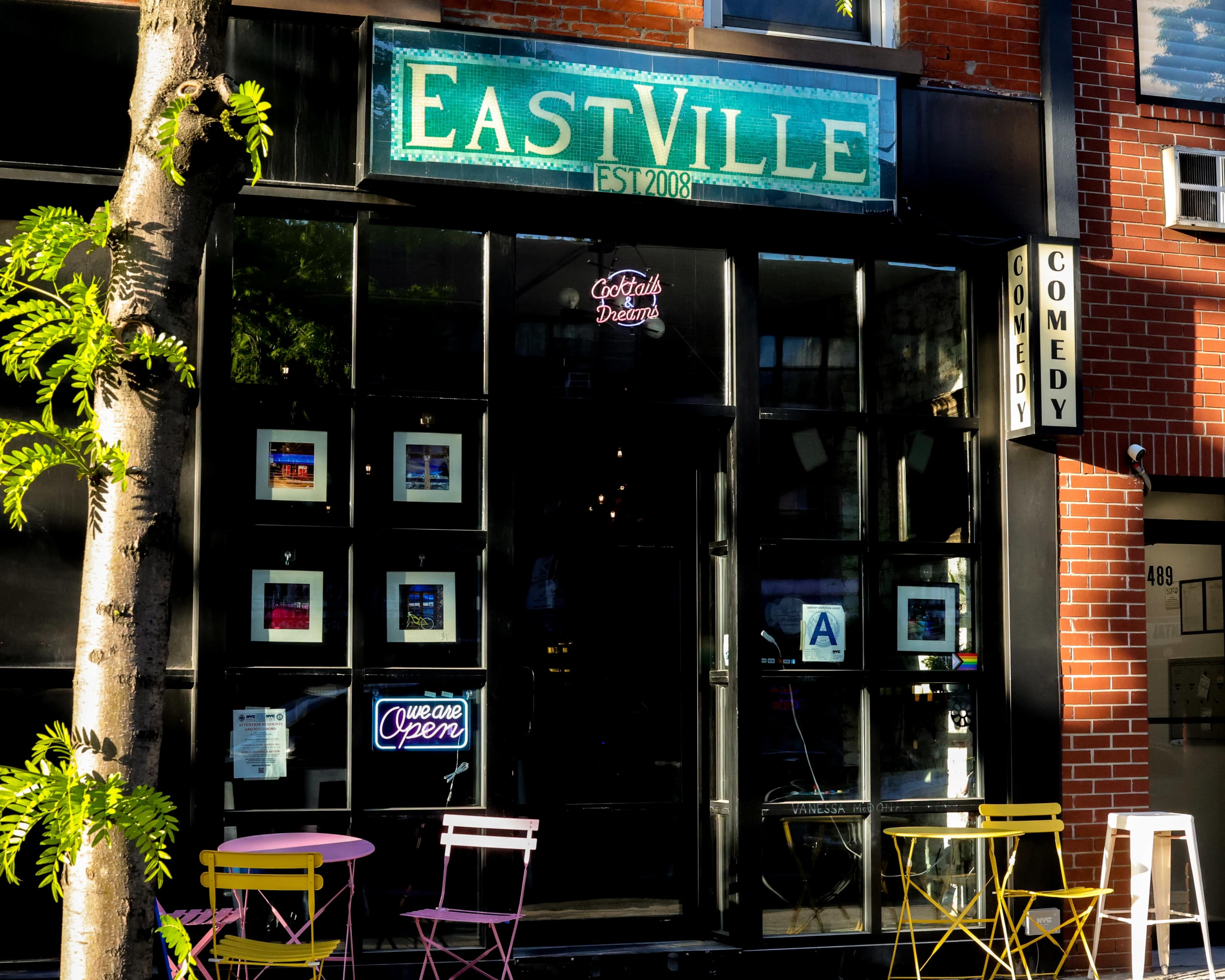 The image shows the exterior of a venue named "EastVille," featuring large glass windows with framed artwork, colorful chairs outside, and signage indicating it's open.