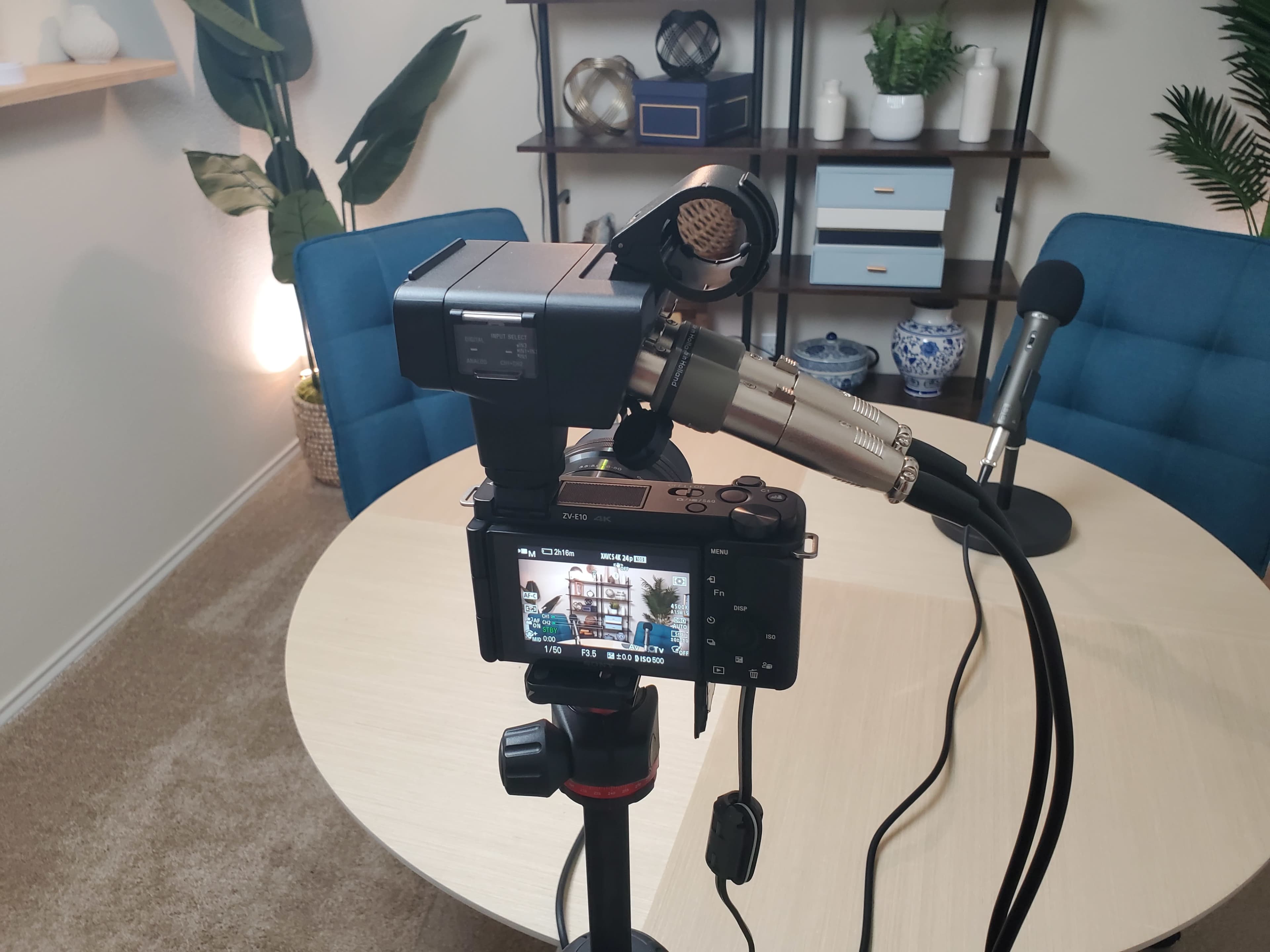 A camera set on a tripod captures a microphone and a table with blue chairs in a well-lit room.