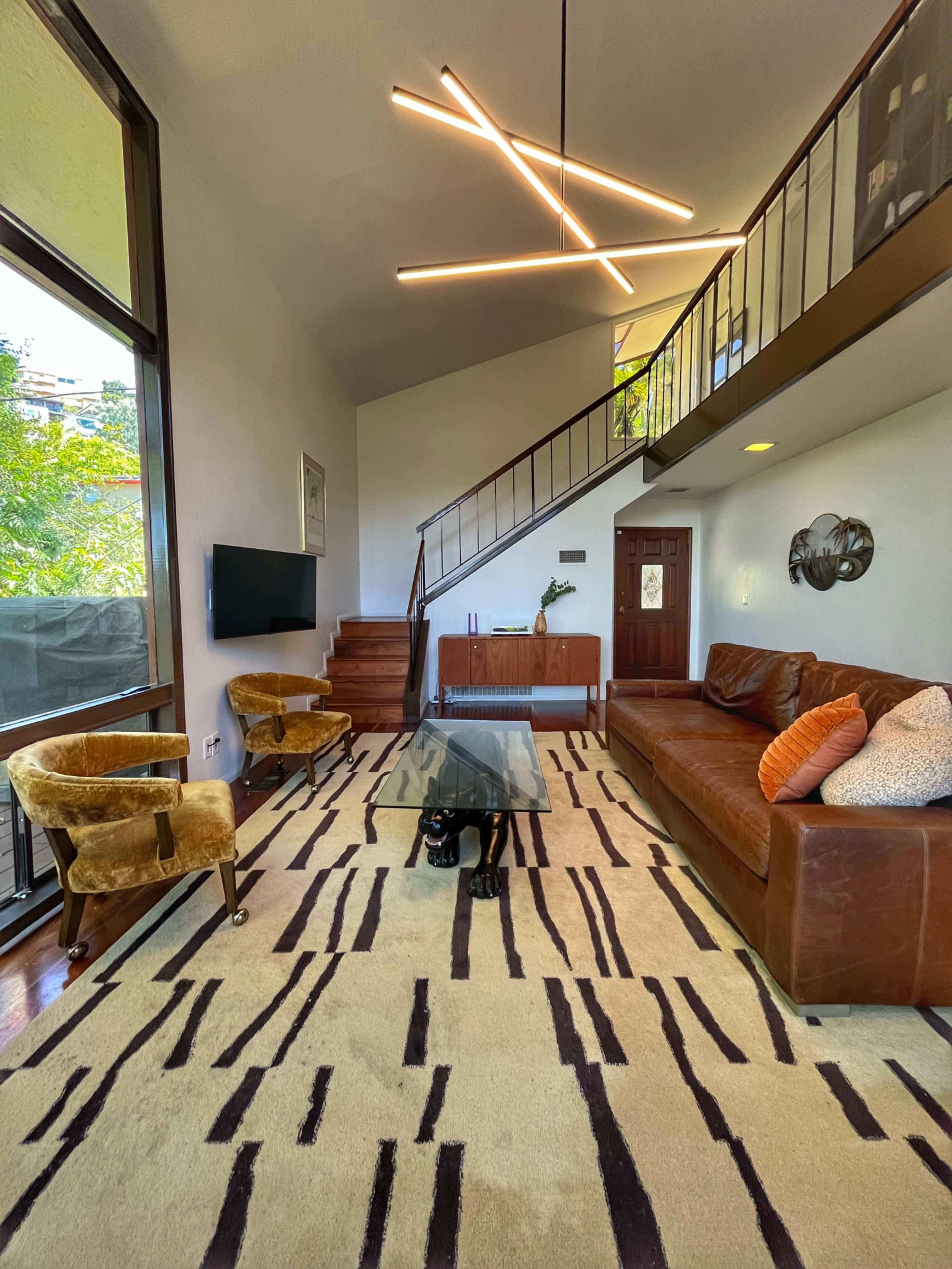 A modern living room features a staircase, leather sofa, glass coffee table, and decorative lighting above a patterned rug.