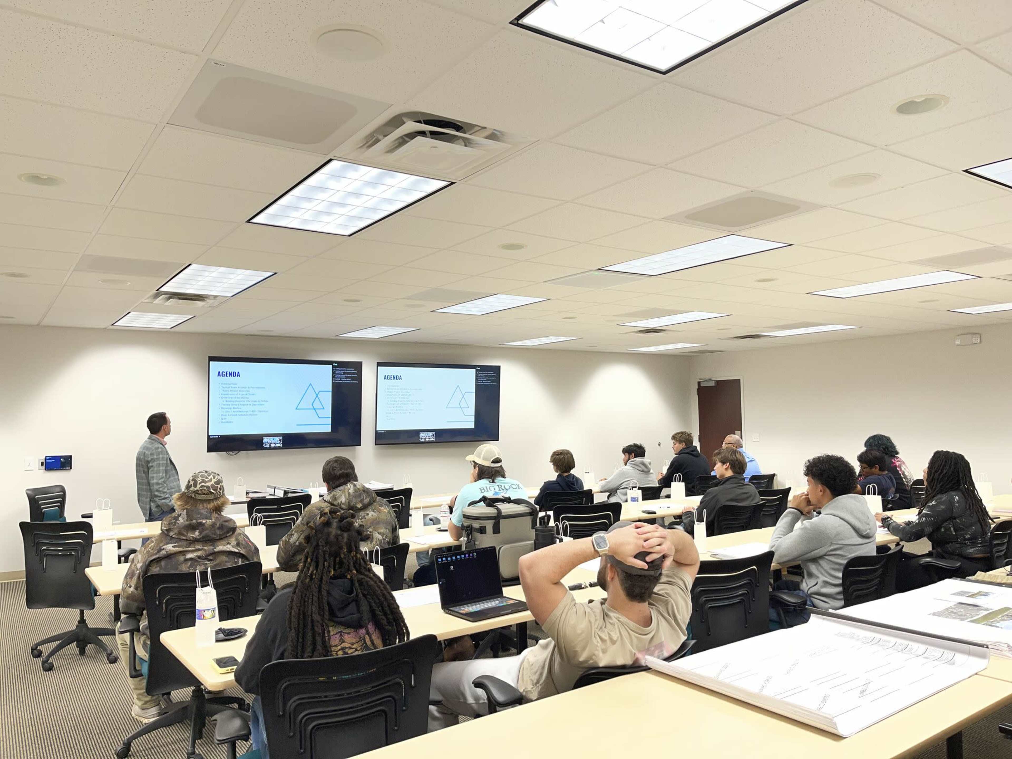A group of students sits in a classroom facing two large screens displaying a presentation, while a teacher stands at the front.
