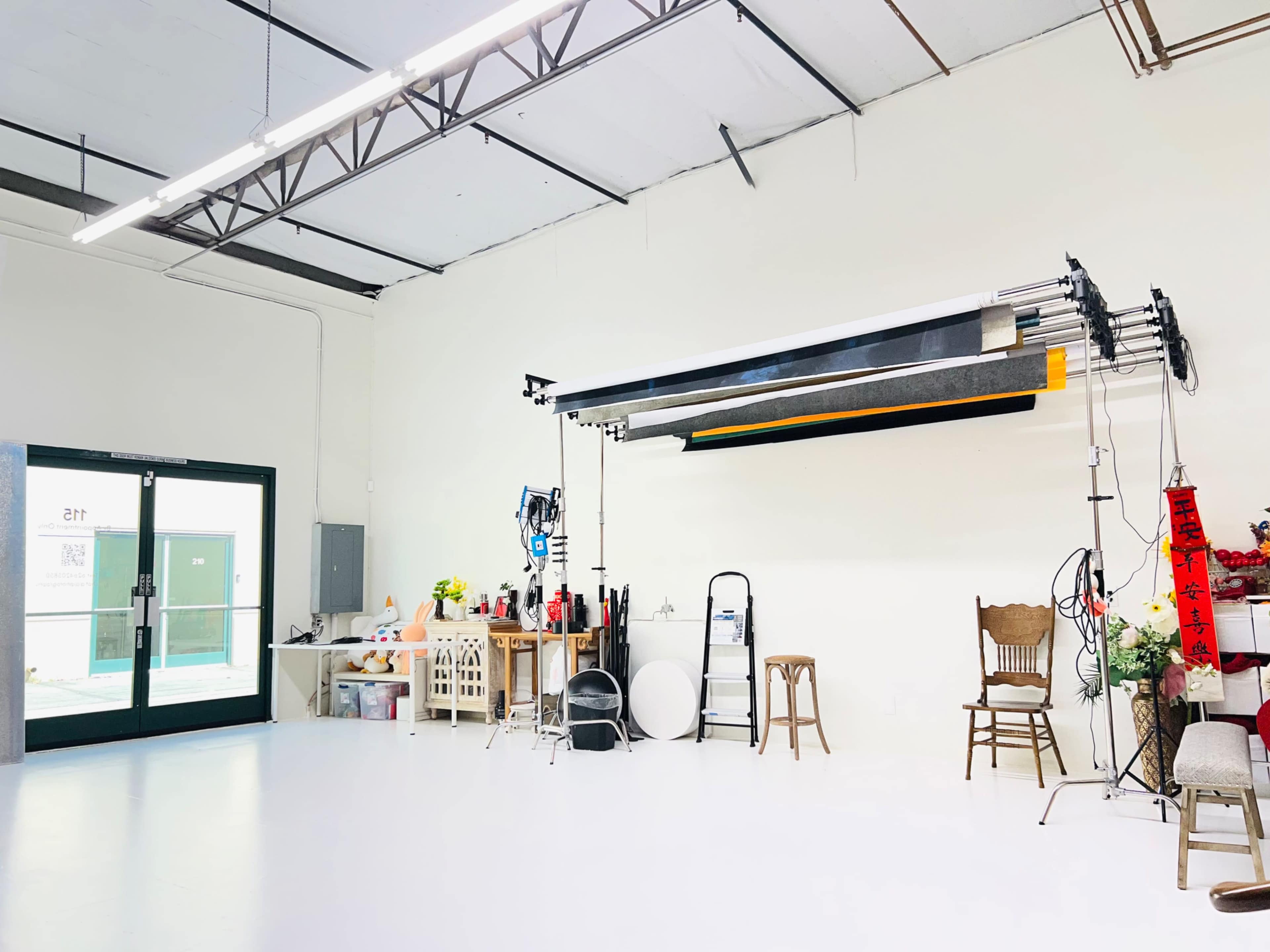 The image shows a spacious studio with white walls, a large open door, various photography equipment, and a few pieces of furniture.