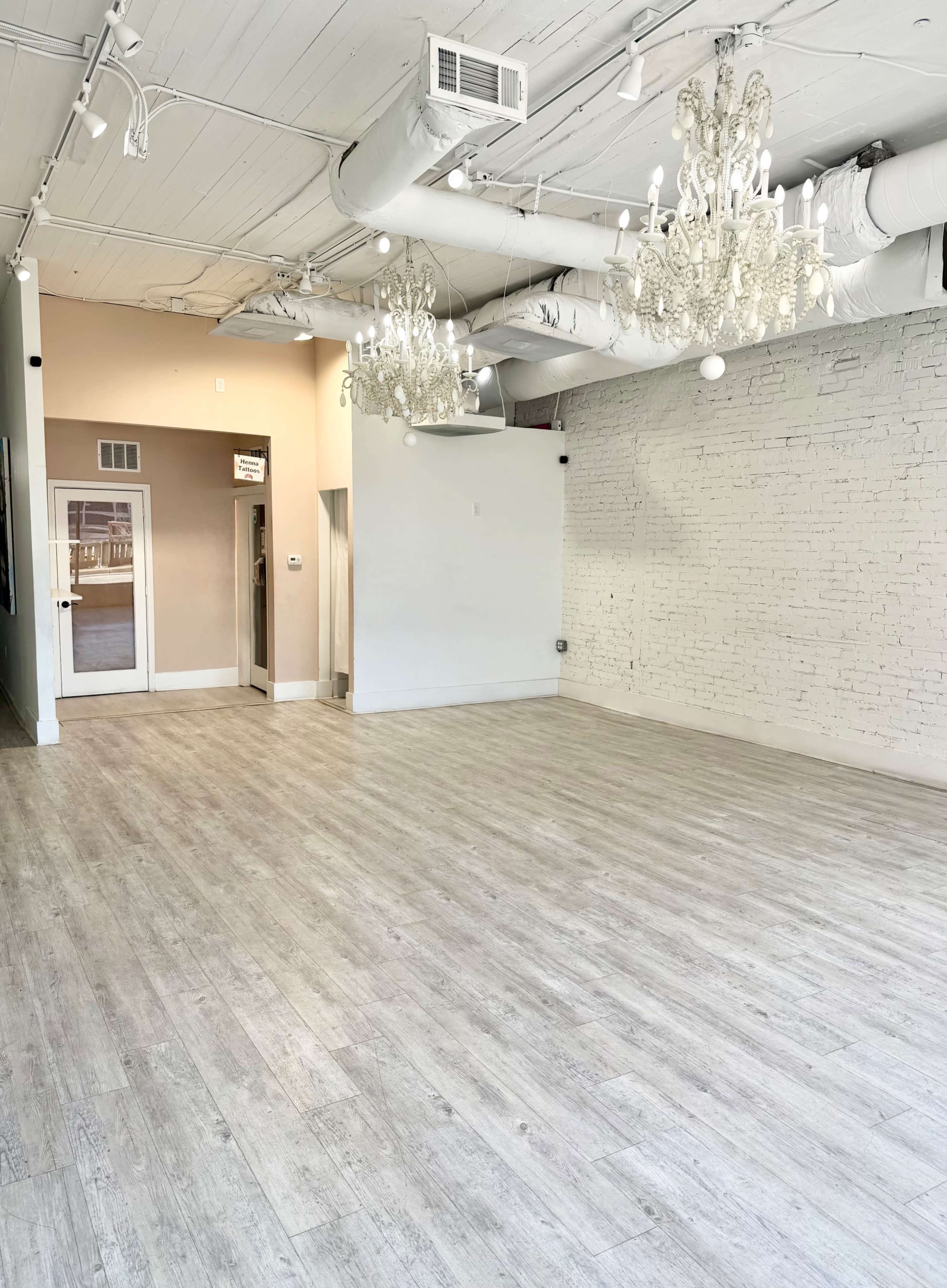 The image shows a spacious, empty room with white walls and a light wooden floor, featuring two chandeliers hanging from the ceiling.
