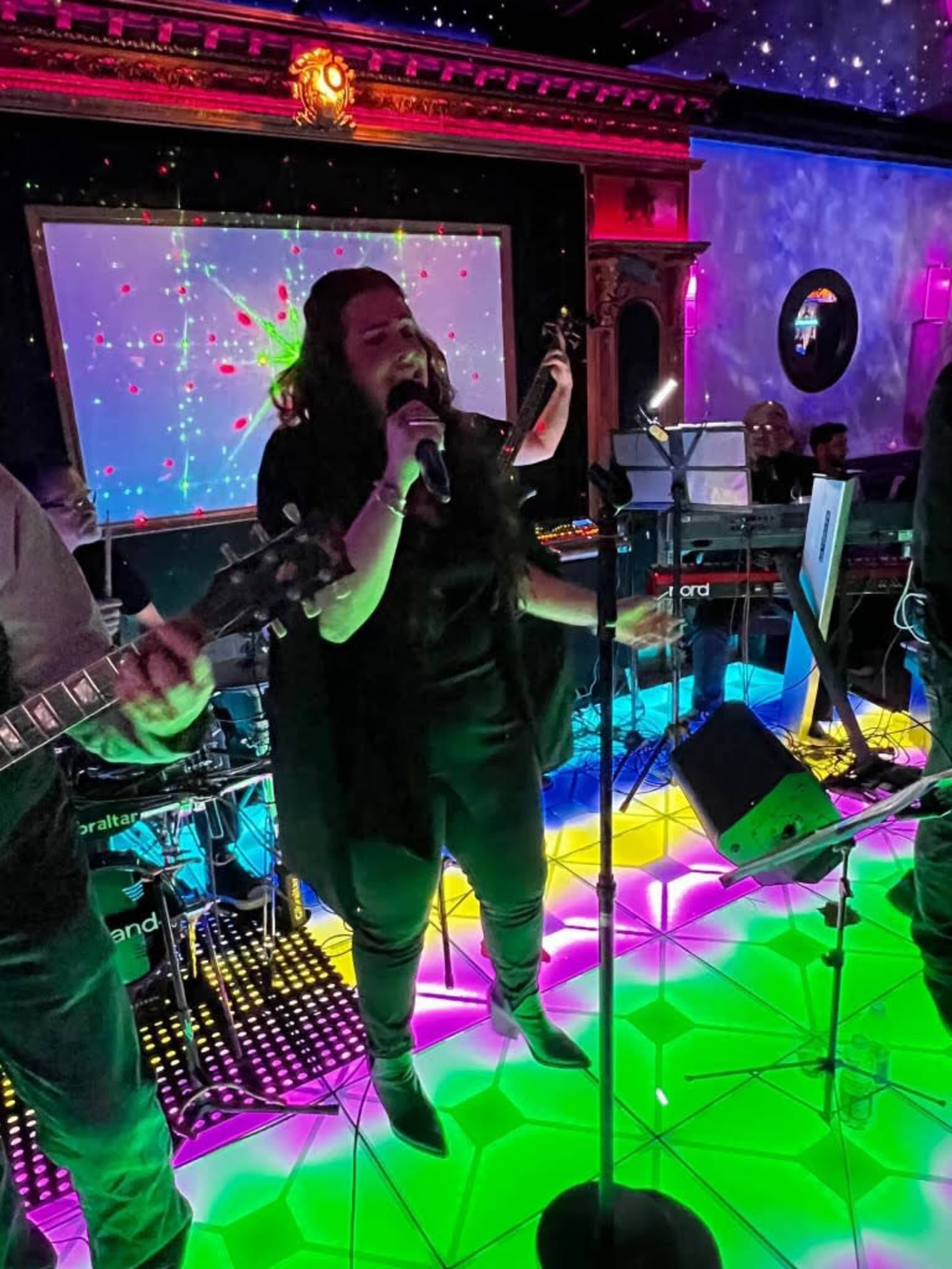 A singer performs on a colorful illuminated stage surrounded by musicians with instruments.