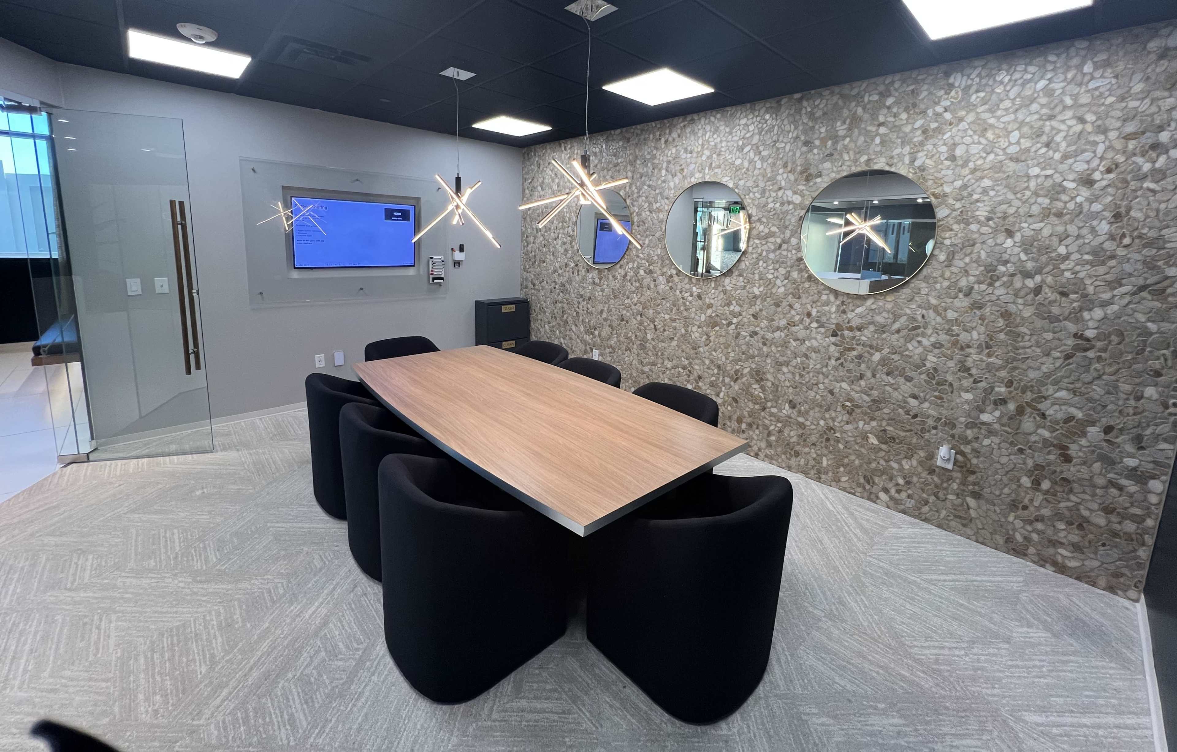 The image shows a modern conference room featuring a large wooden table surrounded by black, rounded chairs, and a stone accent wall with circular mirrors.