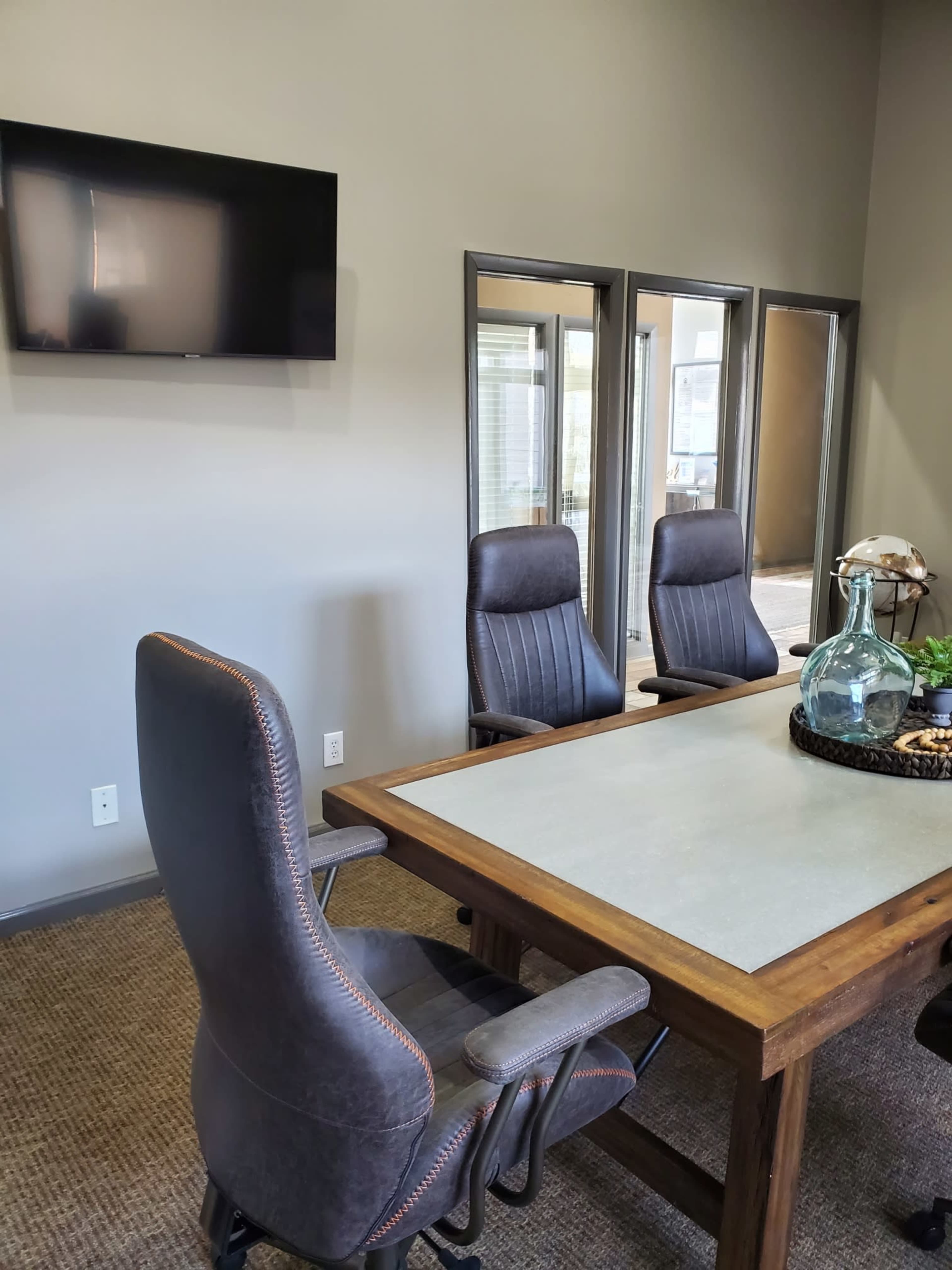 A conference room features a wooden table surrounded by three chairs, with a television mounted on the wall and mirrors reflecting the space.