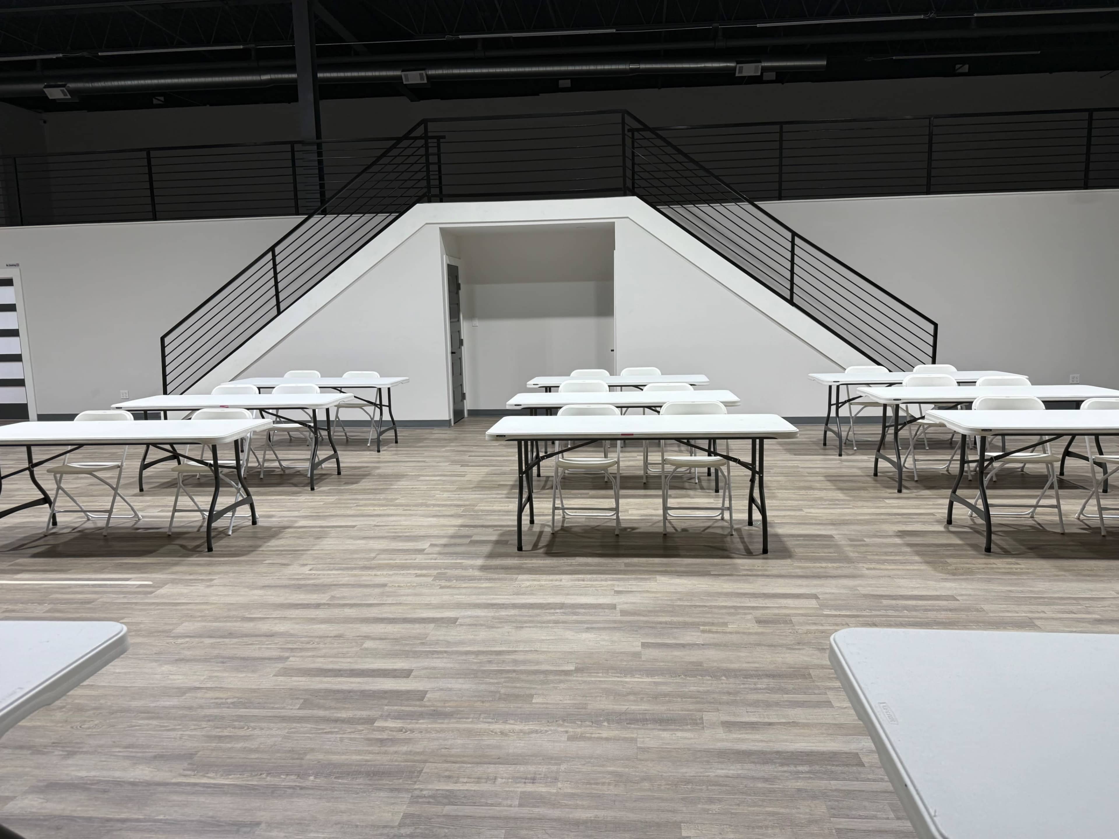 The image shows a spacious, empty room with several white tables set up in rows and a staircase in the background.